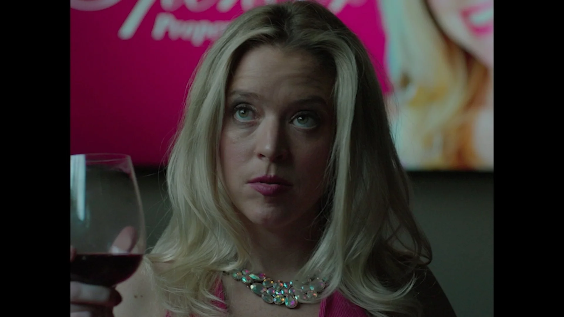 A woman with blonde hair holding a glass of red wine, wearing a colorful necklace and a pink top, sitting at a table in a setting with a pink background featuring partially visible text.