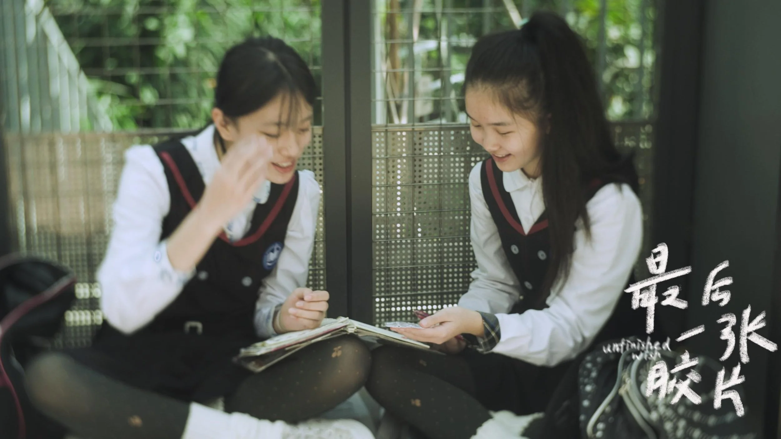 Two schoolgirls in uniforms sitting on the ground, looking at a booklet and smiling, with a metal fence and greenery in the background.
