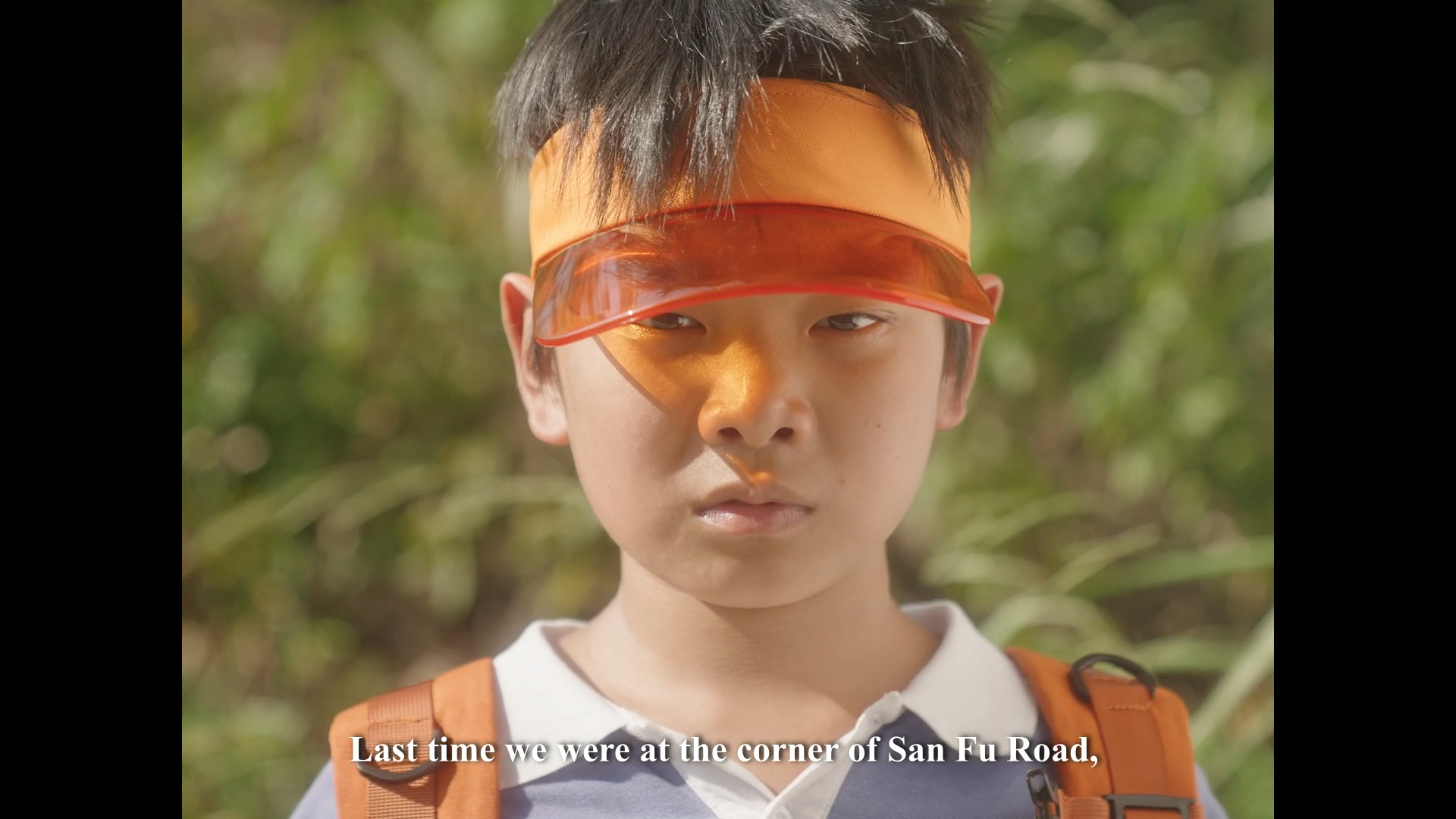A young boy with short dark hair wearing an orange visor, white collared shirt, and orange backpack, standing outdoors with sunlight and greenery in the background.