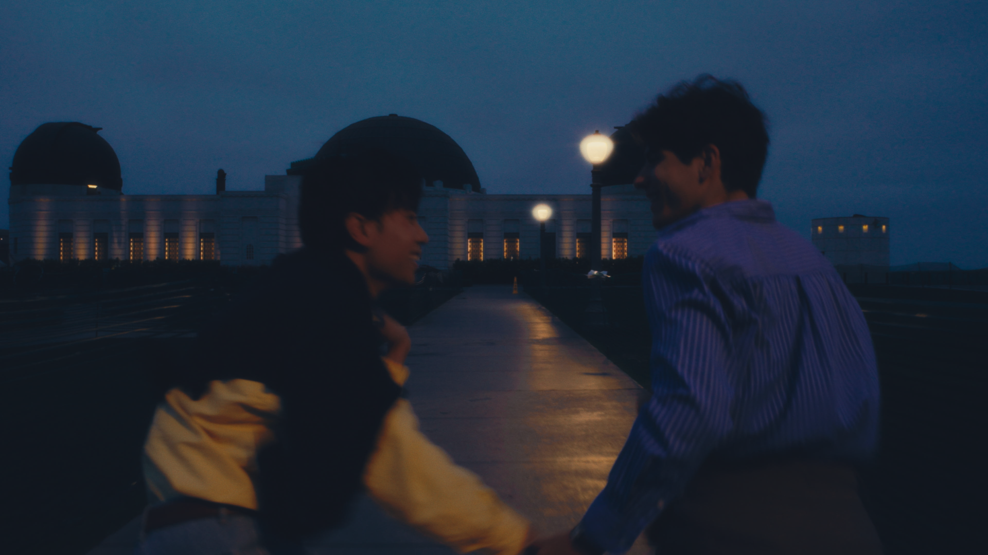 Two young men holding hands and smiling at each other outdoors during evening, with the Griffith Observatory illuminated in the background.