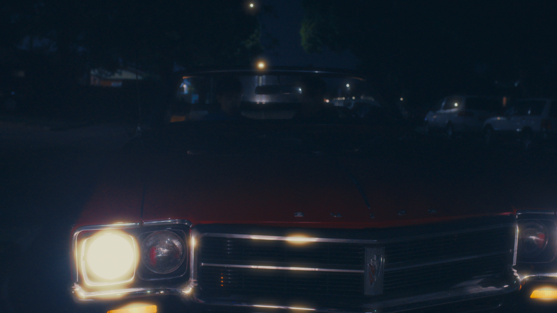 A close-up of the front of a vintage car at night, showing its illuminated headlights, grille, and part of the hood, with parked cars and trees in the background under a moonlit sky.
