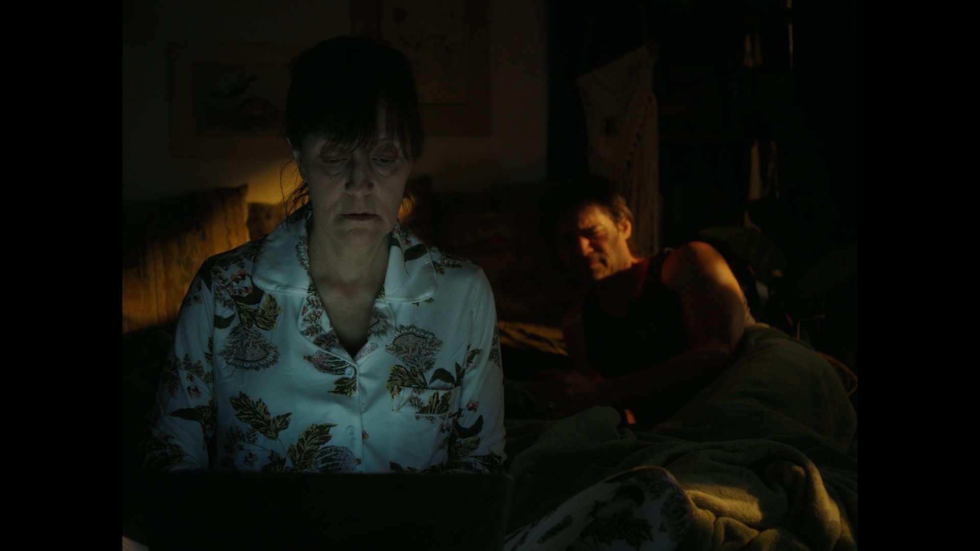 A woman in a floral shirt looks down at something in front of her in a dark room, with a man lying on a bed in the background, appearing to be in pain or discomfort.