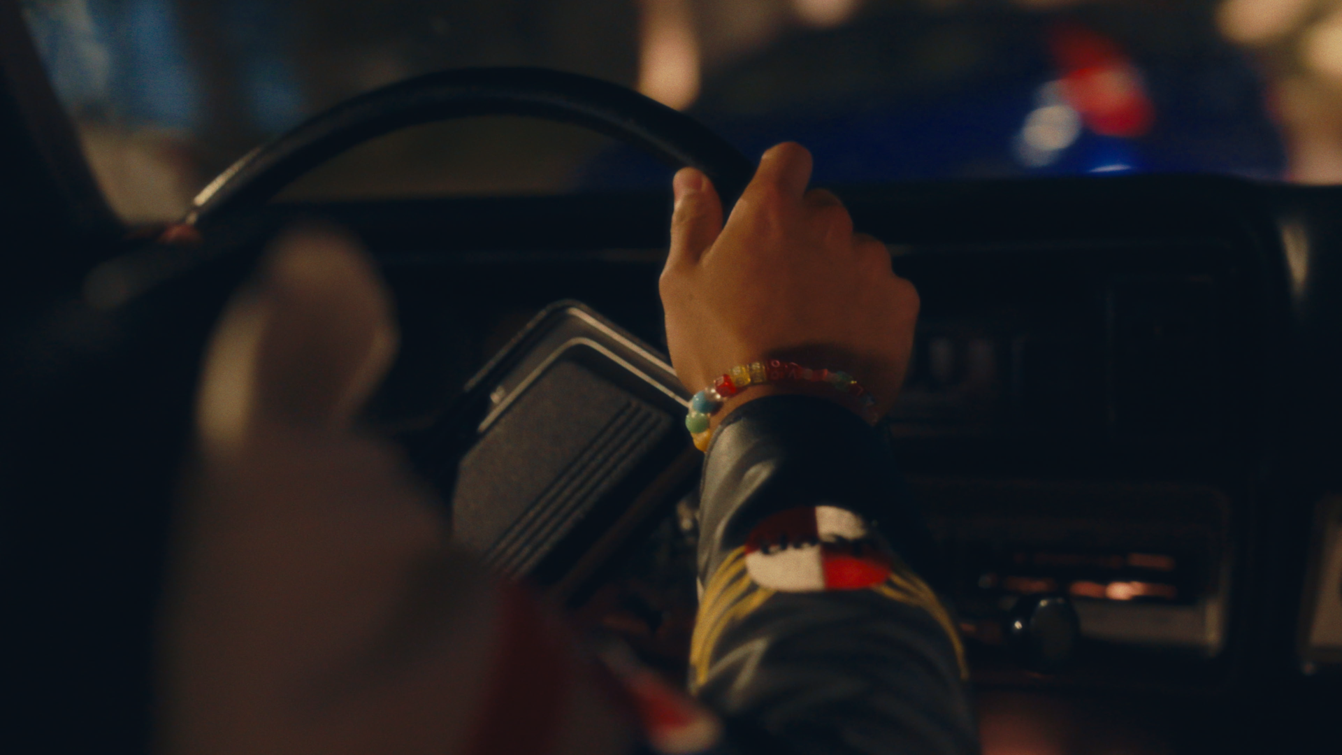 A person driving a car at night, with a focus on their hand gripping the steering wheel. The person is wearing colorful beaded bracelets.