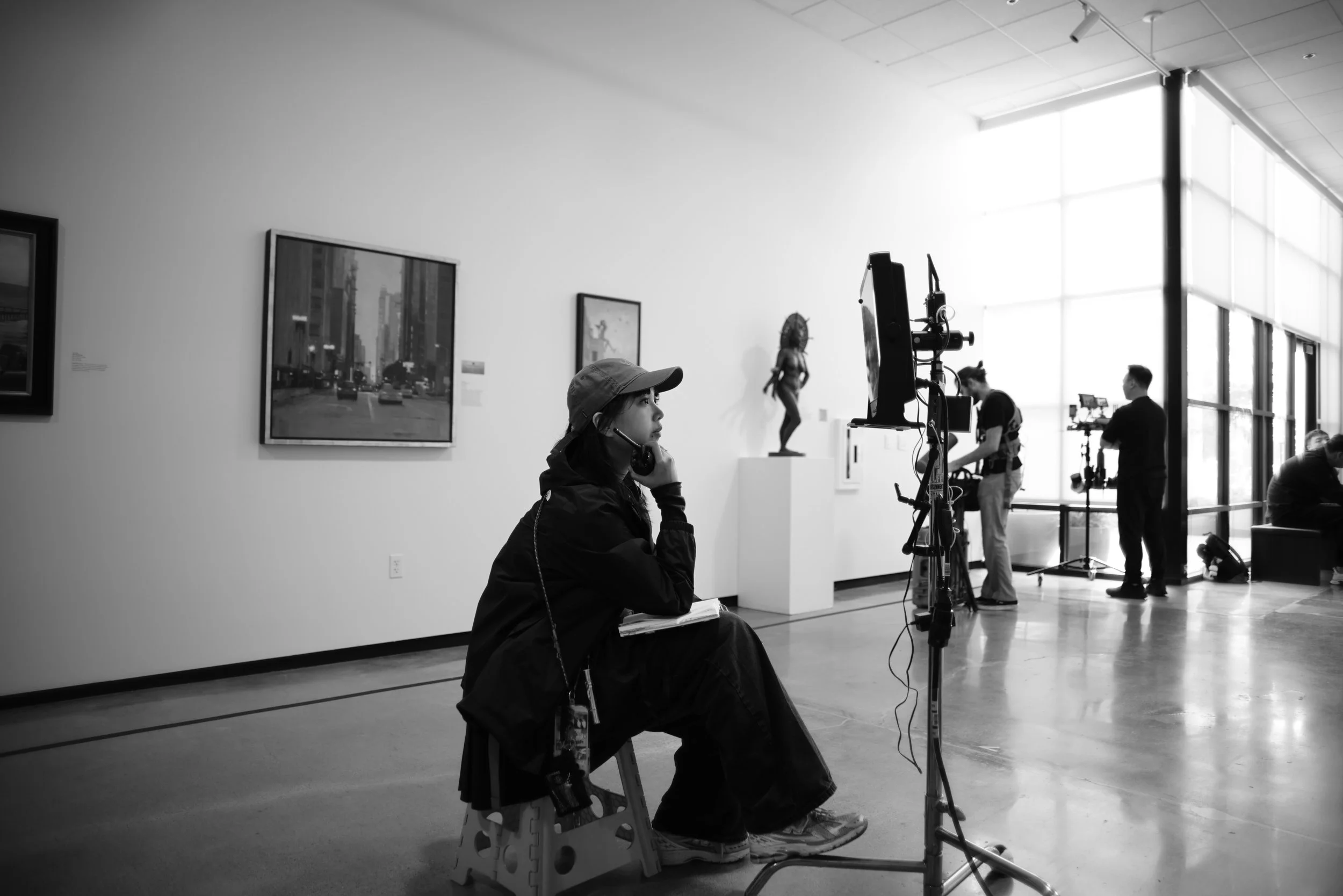 A woman with a cap sitting on a stool in an art gallery, talking on a phone. Artworks hang on the wall behind her, and people are working at equipment in the background.
