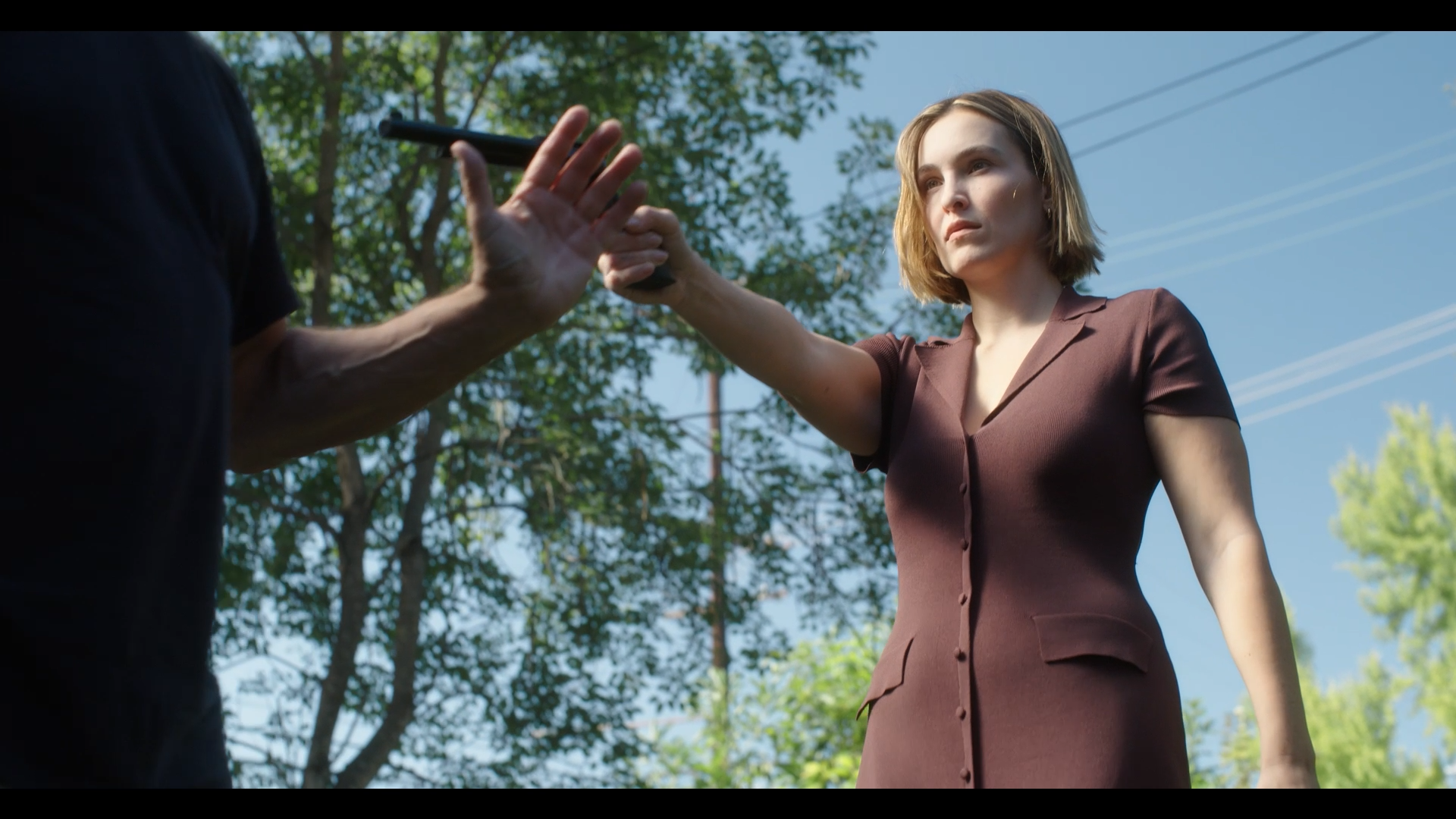 A woman with short hair, wearing a brown button-up dress, stands outside with trees and power lines in the background. Someone off-camera is handing her a gun.