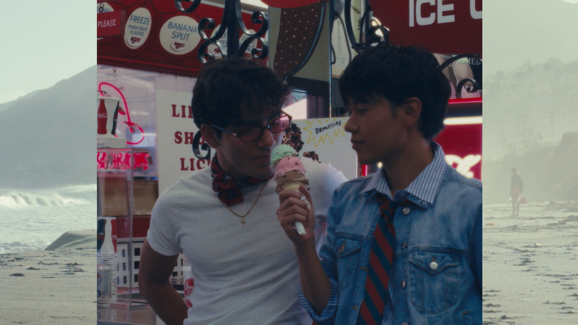 Two young men at an ice cream shop, one holding a three-scoop ice cream cone, with colorful signs in the background.