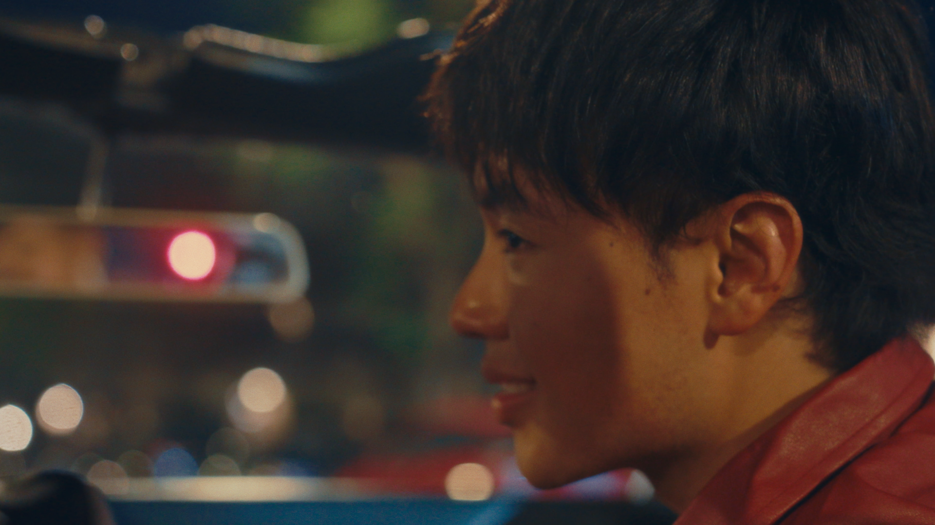 A person with short dark hair, wearing a red jacket, smiling, with blurred city lights in the background.
