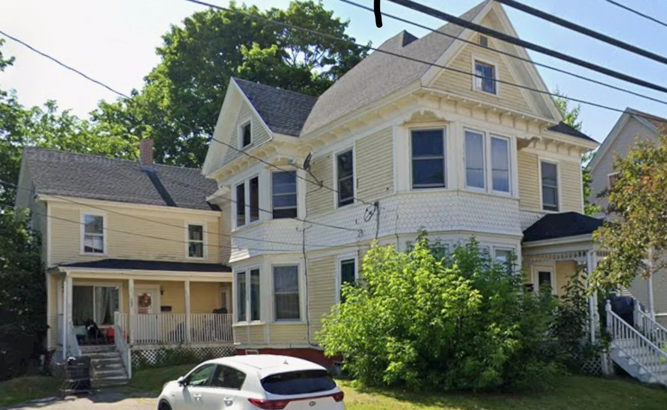 A large yellow Victorian-style house with multiple stories, bay windows, decorative trim, and a front porch. There is a white car parked in front and wires overhead. Lush green trees surround the house.