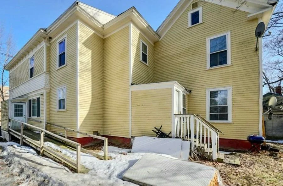 Yellow two-story house with multiple windows, a small deck with stairs, and a ramp, in winter with snow on the ground.