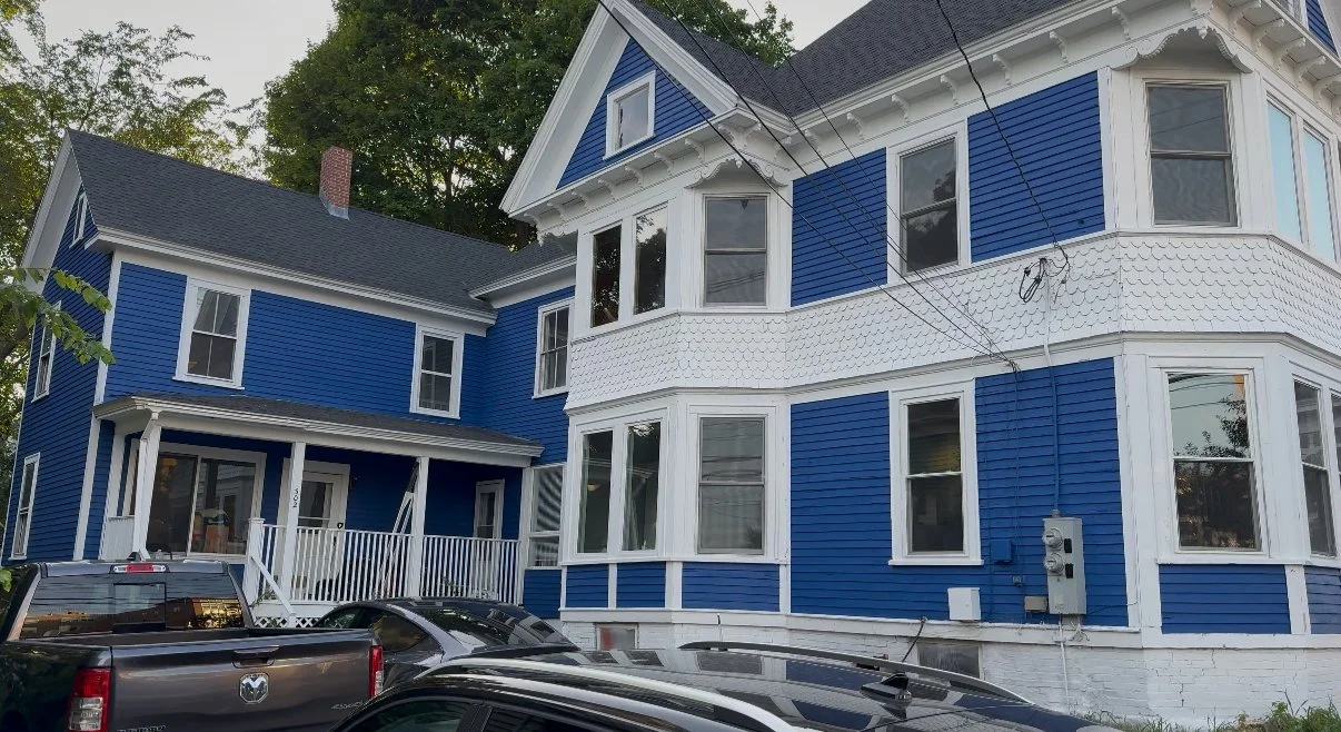 Blue Victorian-style house with multiple windows and a small front porch, cars parked in front, surrounded by trees.