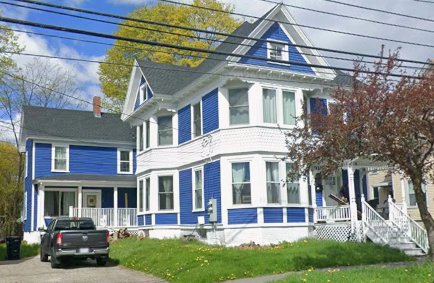 A large two-story blue house with white trim and a wrap-around porch, a black pickup truck parked in the driveway, and a tree with reddish leaves in the front yard, under a partly cloudy sky.