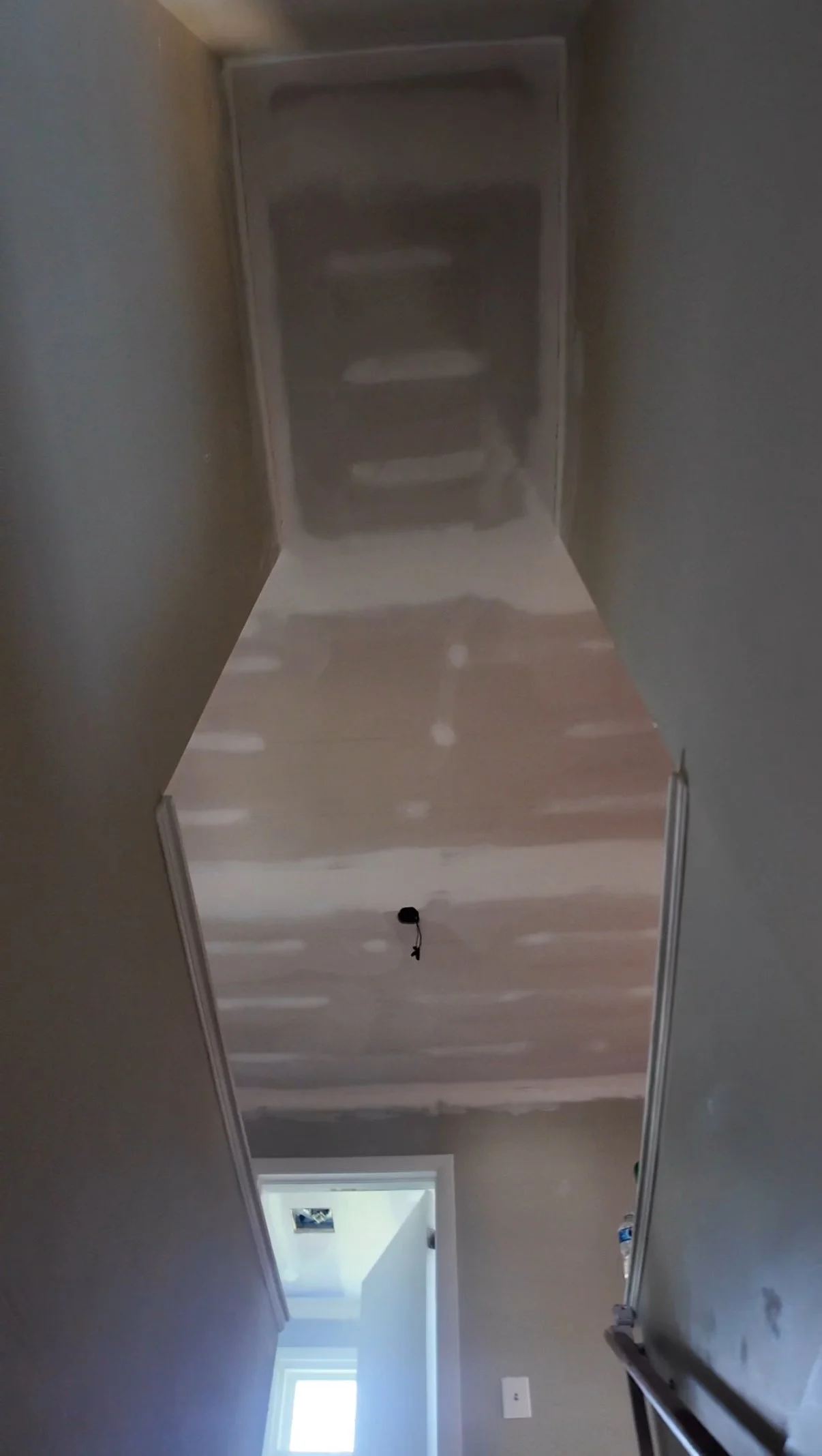 A hallway under construction with unfinished drywall on the ceiling and walls, and a window at the end providing natural light.