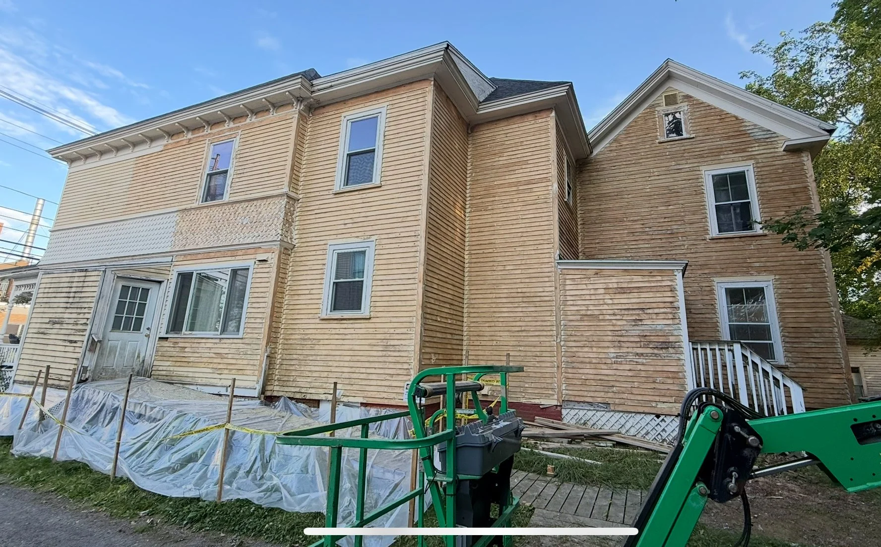 A two-story house undergoing exterior renovation, with part of the siding removed and construction equipment present on the yard.