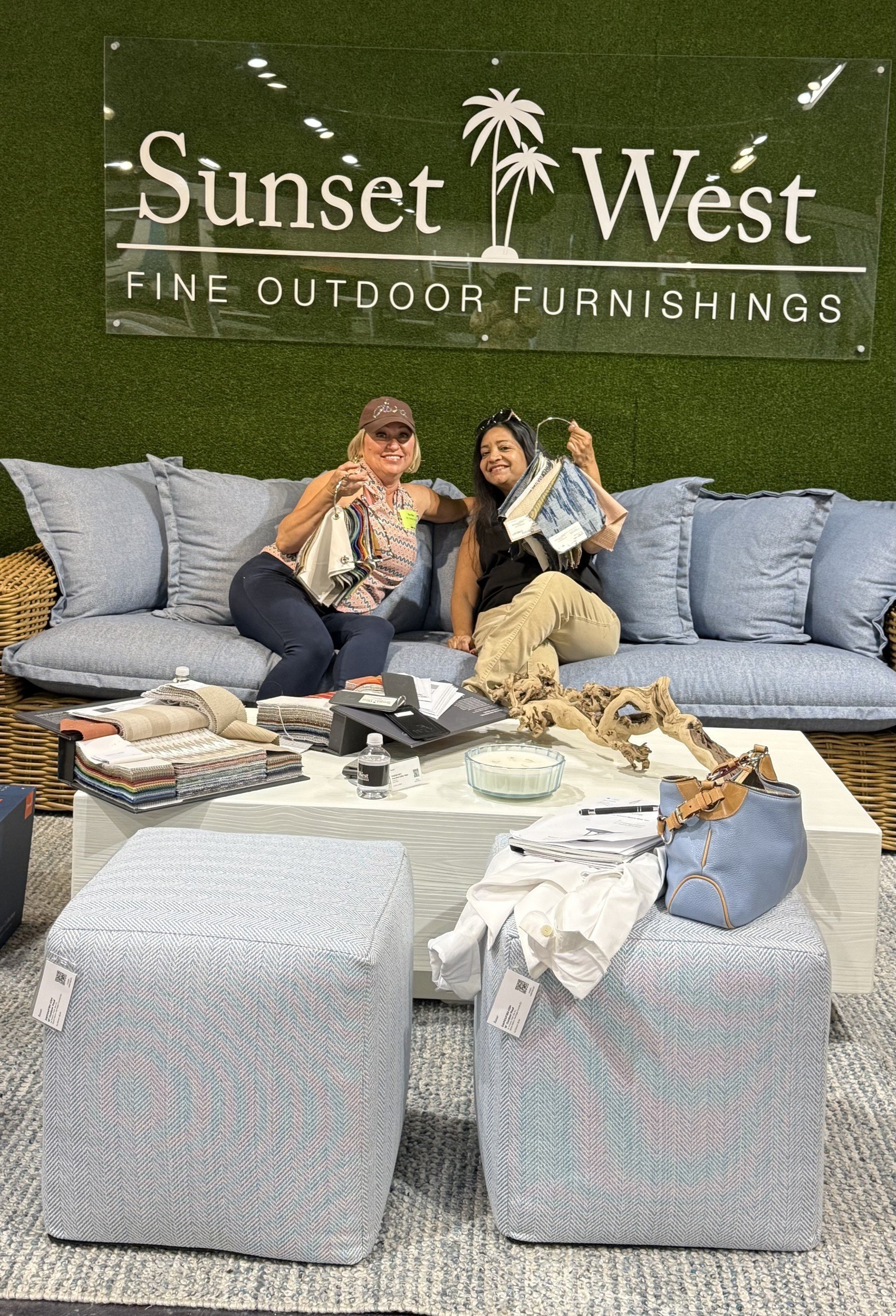 Two women sitting on a gray outdoor sofa holding fabric swatches and smiling. The background features a large sign that reads 'Sunset West Fine Outdoor Furnishings' with a palm tree graphic. In the foreground, there is a white table with fabric samples, a water bottle, a purse, and other items.