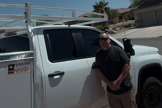 A man standing next to a white pickup truck with a ladder rack on top, parked in a suburban neighborhood with palm trees and houses in the background.