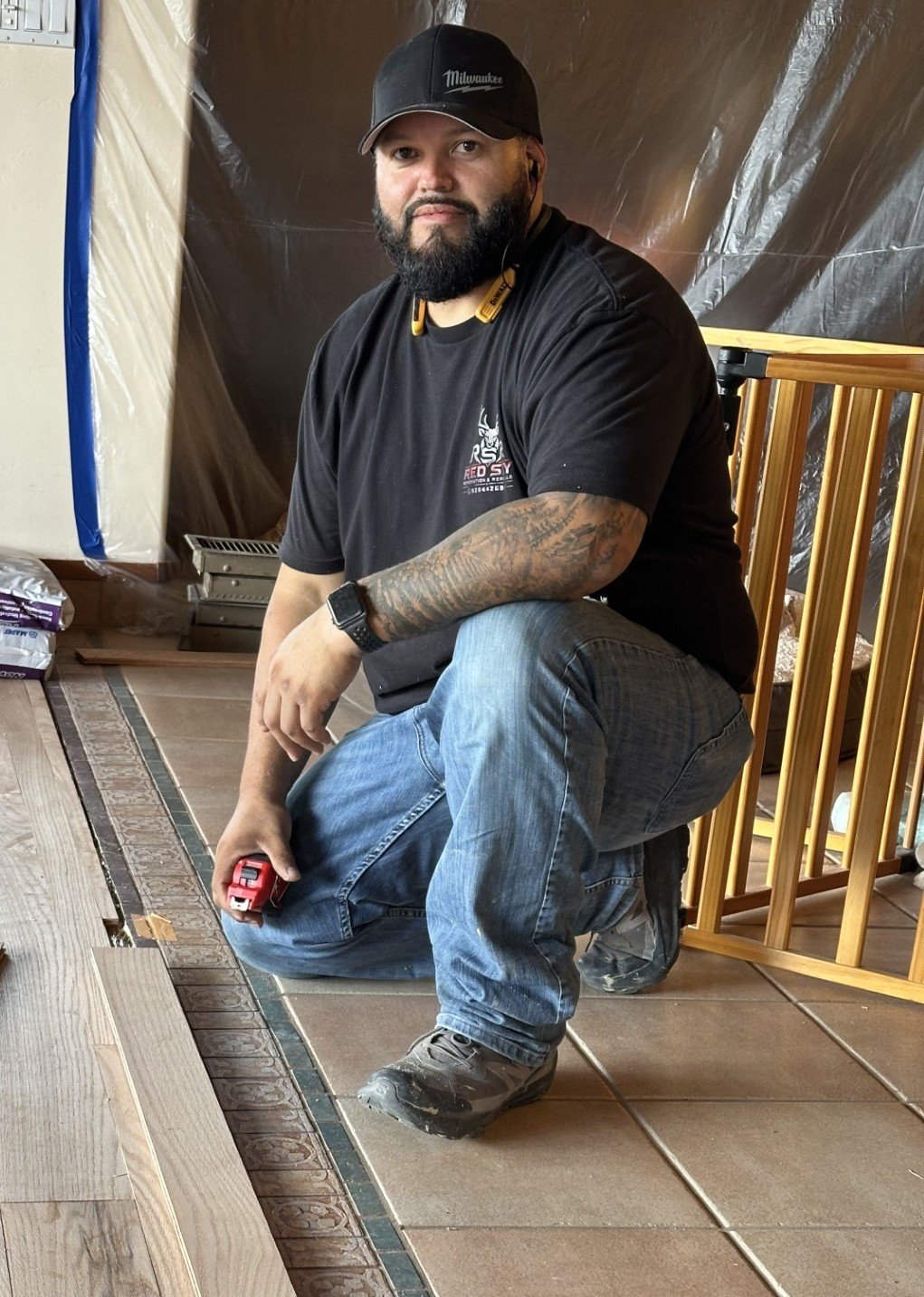 A man with a beard, black cap, and black t-shirt kneeling on a tiled floor, holding a measuring tape, and working on flooring installation.