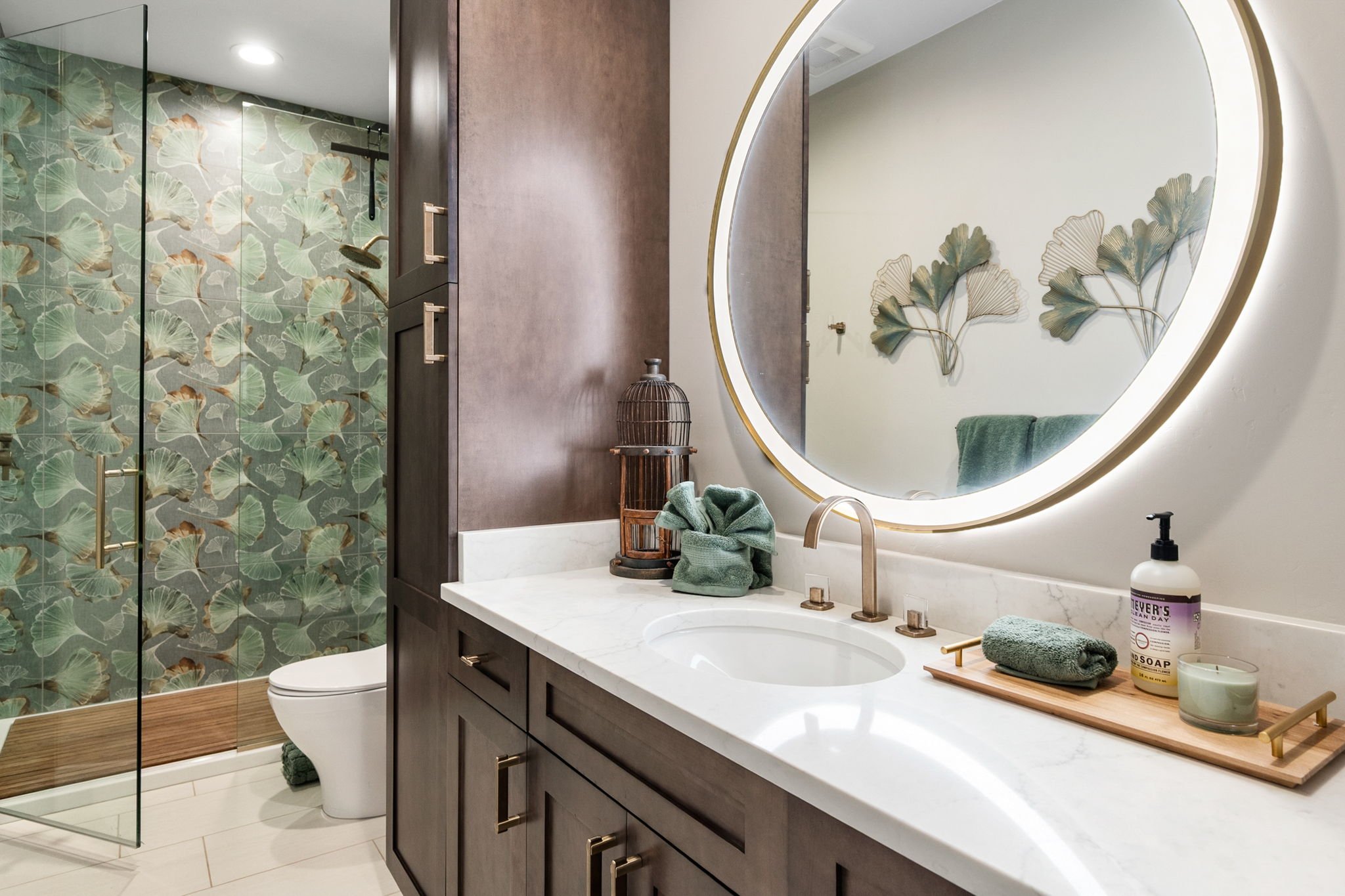 Refined guest bathroom featuring a rich wood vanity, illuminated round mirror, botanical accents, and a statement glass-enclosed shower with softly patterned tile.