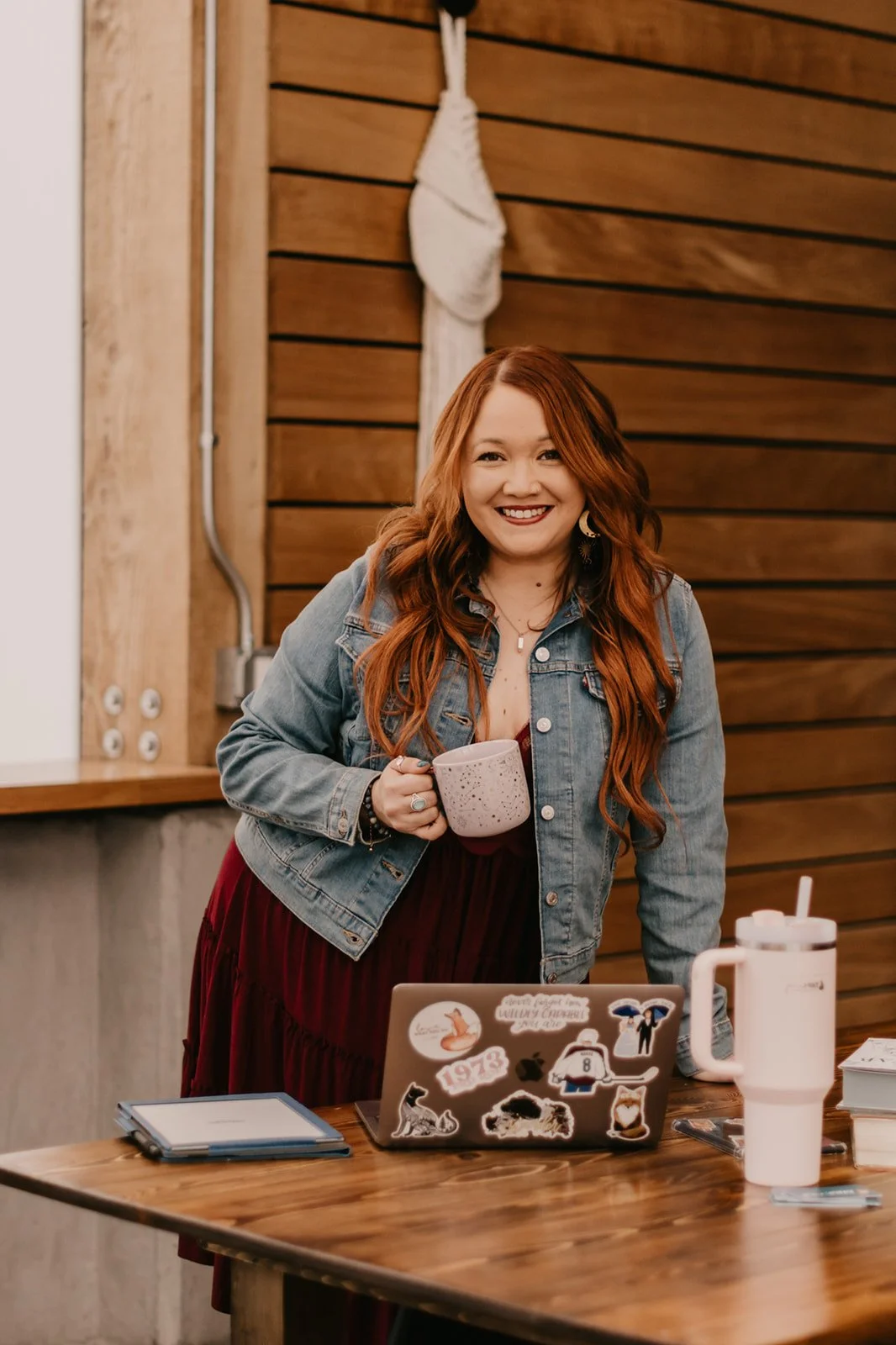 A woman with long red hair, wearing a denim jacket and red dress, smiling and holding a mug in a cozy coffee shop with wooden walls, a laptop with stickers, and a reusable drink cup on a wooden table.