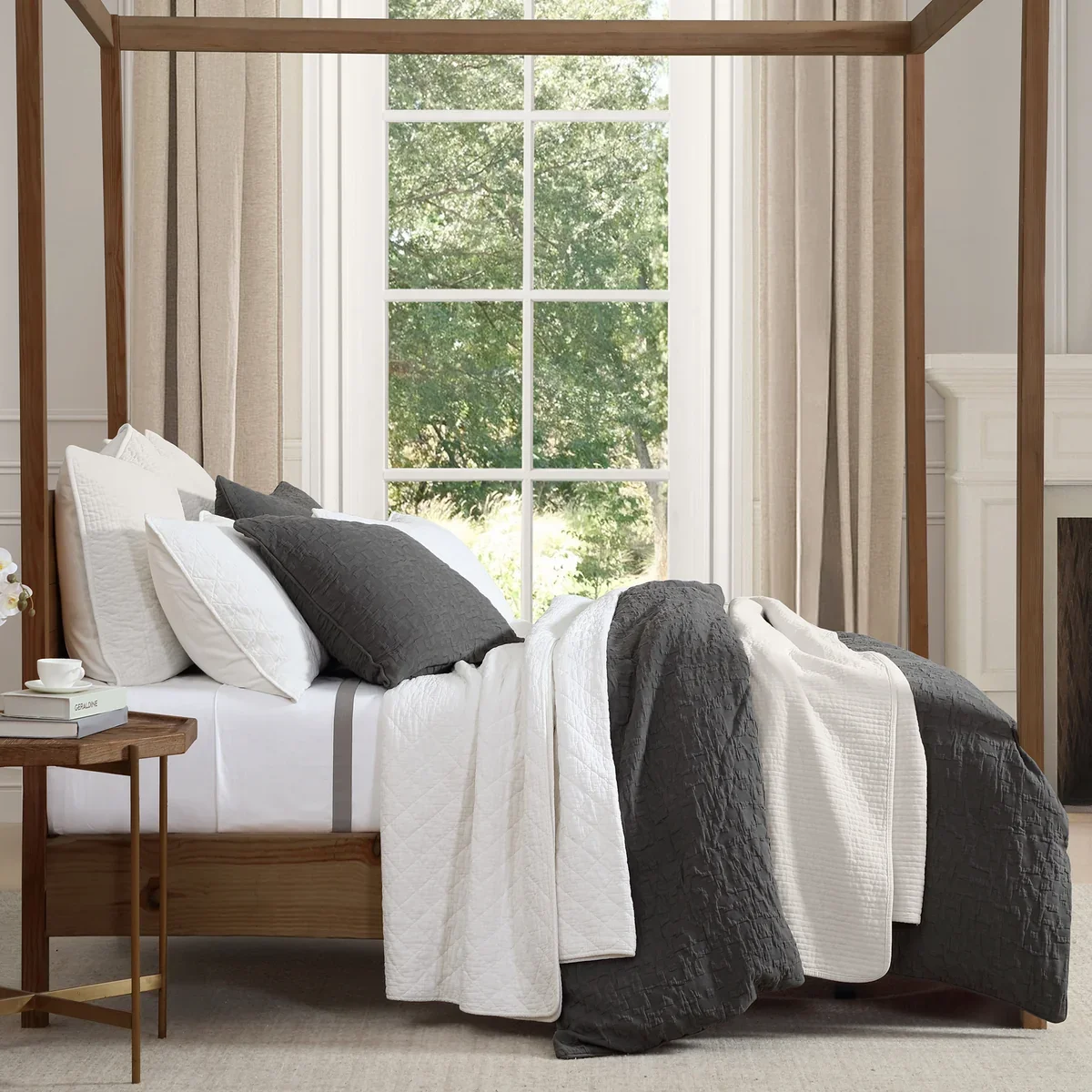 A bedroom with a wooden bed frame, black and white bedding, and large window with beige curtains showing green trees outside.