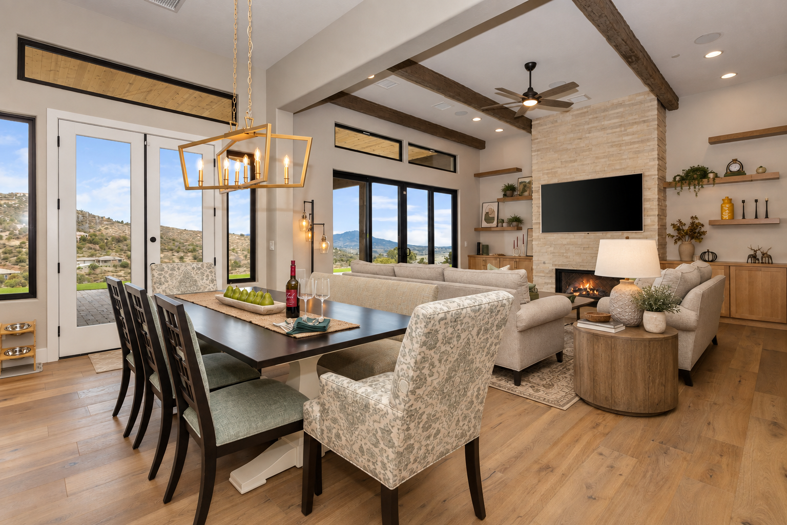 Open living and dining room with large windows showcasing mountain views, a dining table with chairs, a sectional sofa, a fireplace with a wall-mounted TV, and decorative shelves with plants and ornaments.