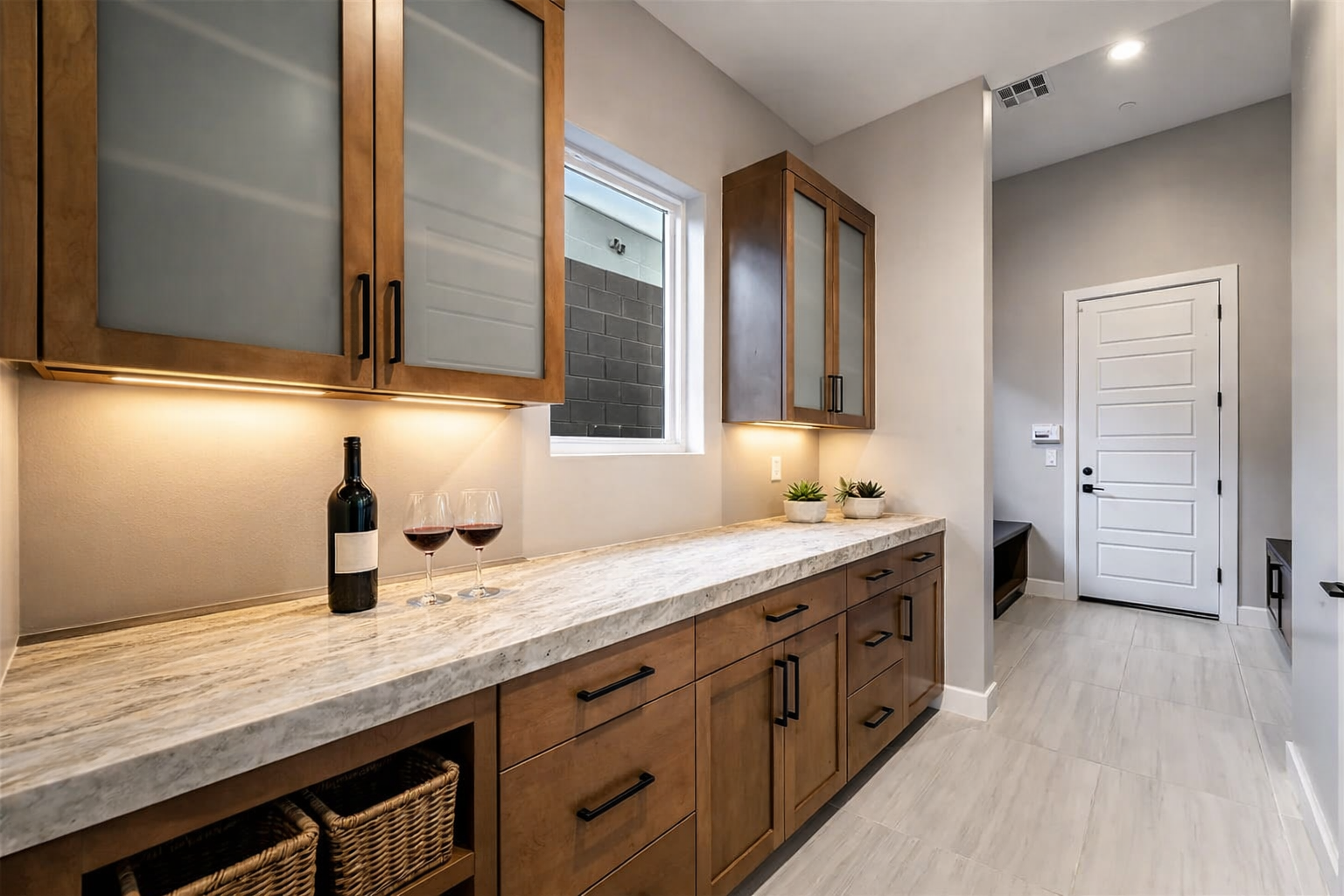 Butler’s pantry and mudroom featuring warm wood cabinetry, glass-front uppers, stone countertops, under-cabinet lighting, and integrated storage.