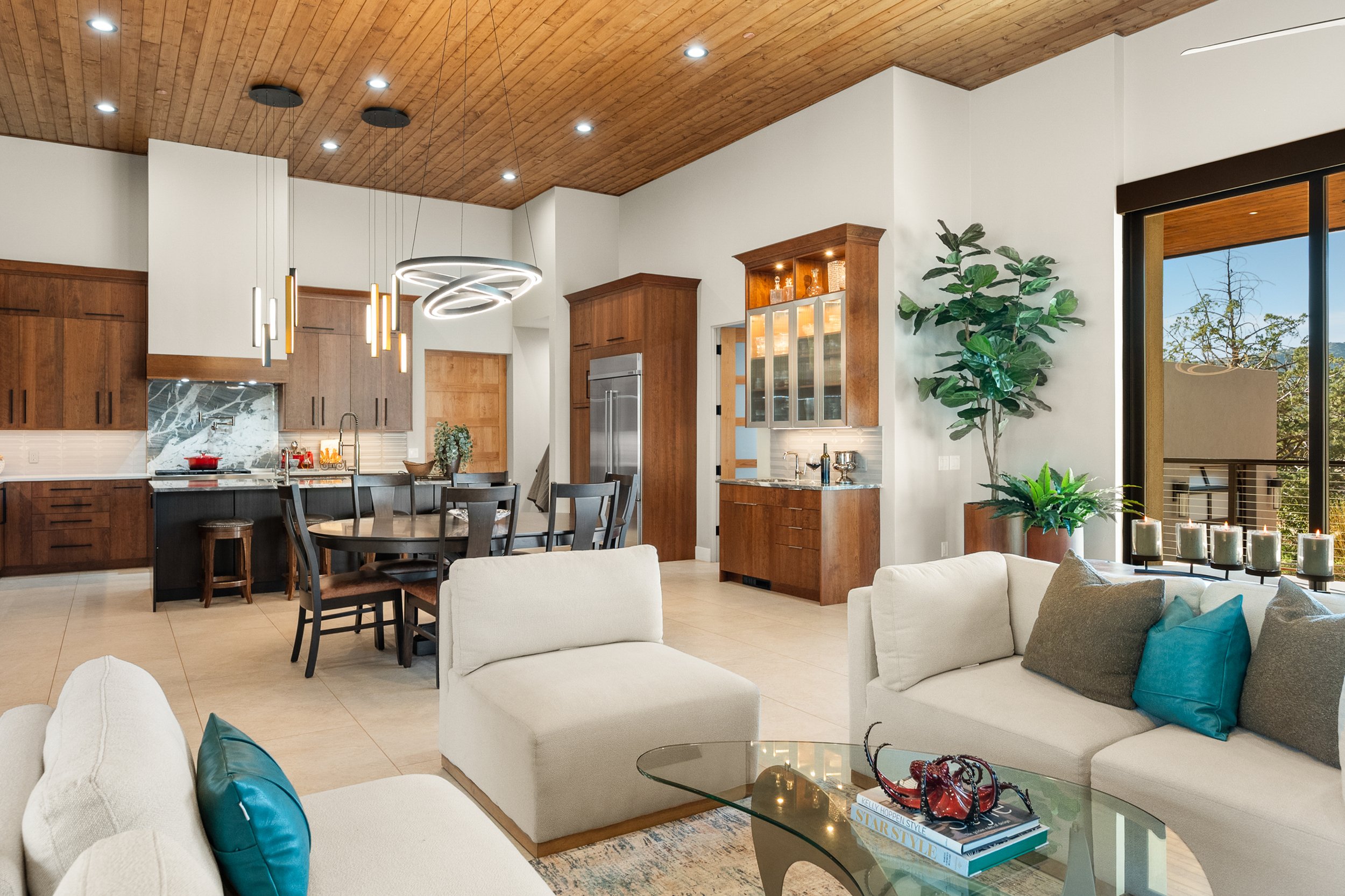 Open-concept living and dining space featuring warm wood ceilings, custom cabinetry, expansive windows, and layered contemporary furnishings.
