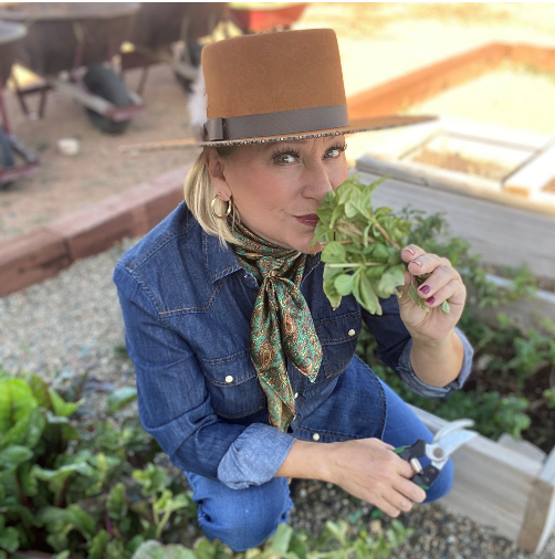 Woman in a blue denim jacket and wide-brimmed hat holding a bunch of fresh green herbs near her face, in a garden setting.