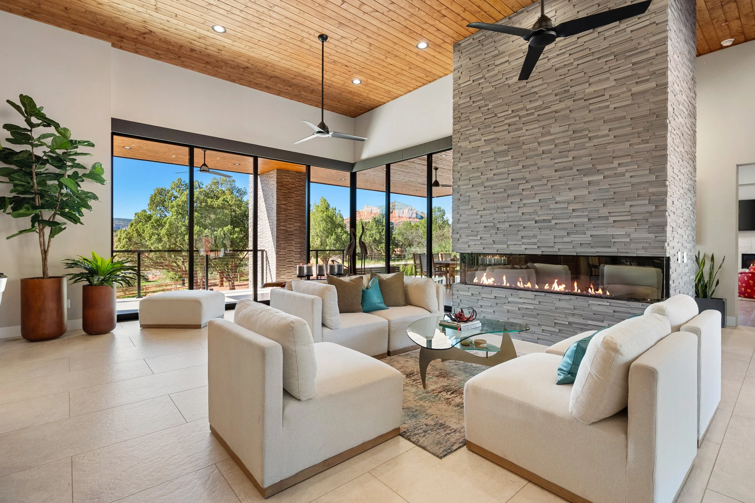 Light-filled great room with floor-to-ceiling windows, a dramatic stone fireplace, warm wood ceiling, and layered contemporary furnishings.