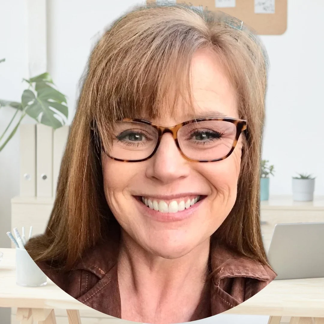 Close-up of a smiling woman with red hair, glasses, and wearing a brown leather jacket, in an office setting.