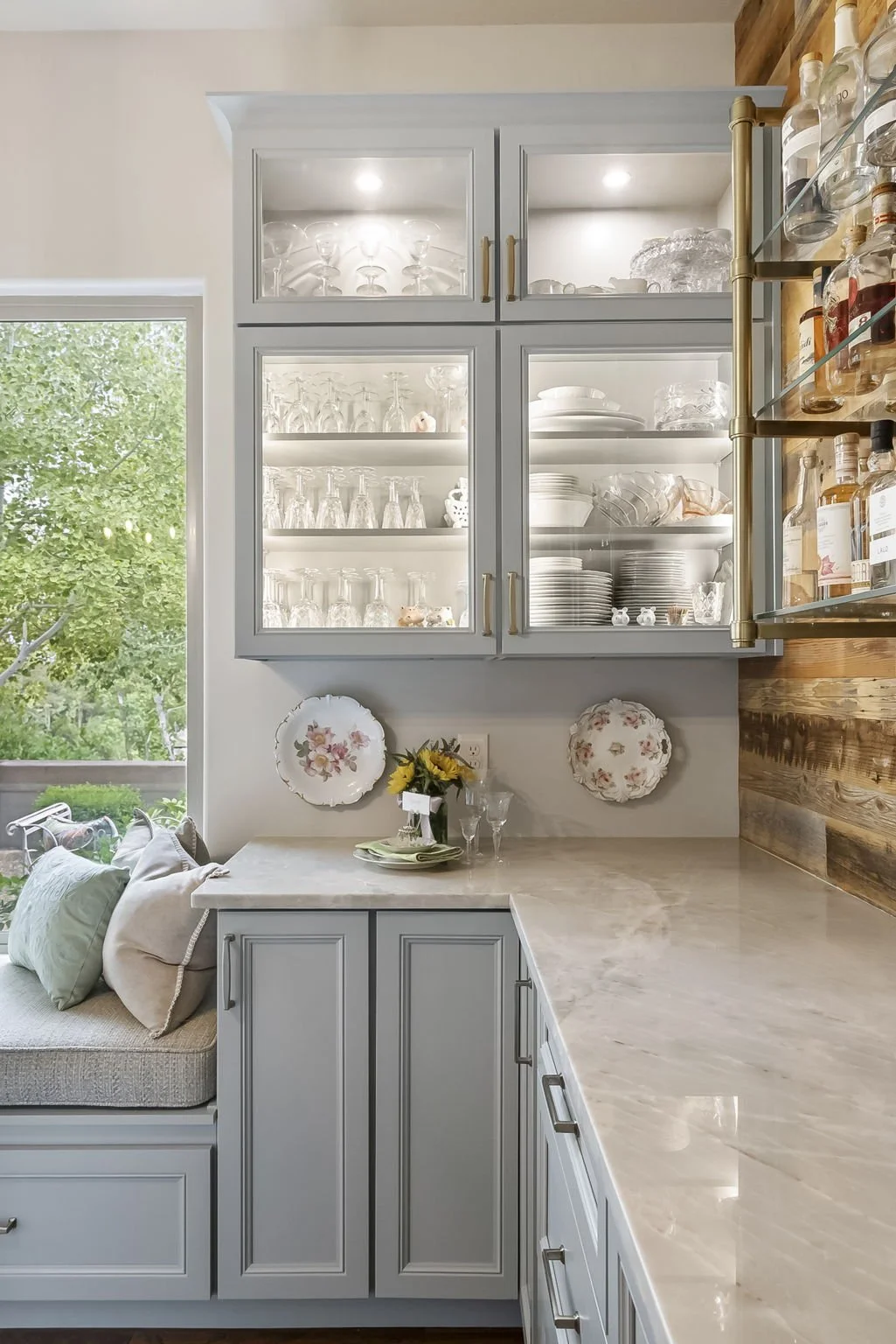 Custom kitchen detail featuring illuminated glass-front cabinetry, quartz countertops, soft gray millwork, and a built-in window seat framed by warm reclaimed wood.