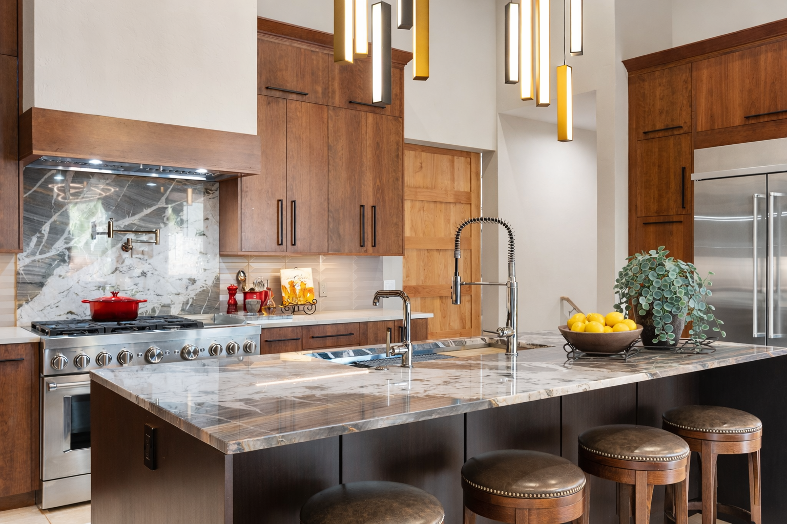 Custom kitchen featuring rich wood cabinetry, dramatic stone surfaces, an oversized waterfall island, sculptural pendant lighting, and generous counter seating designed for gathering.