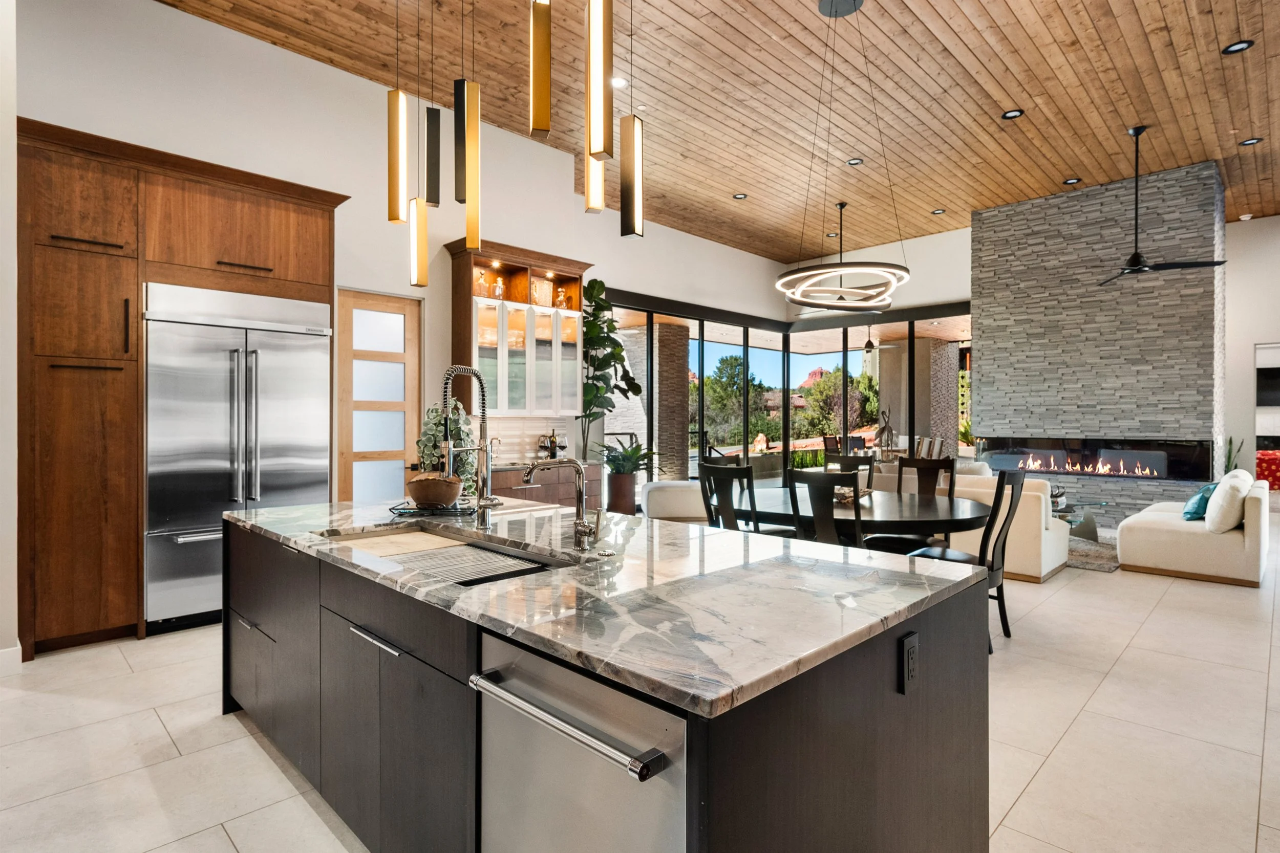 Custom kitchen featuring rich wood cabinetry, dramatic stone surfaces, an oversized waterfall island, sculptural pendant lighting, and generous counter seating designed for gathering.