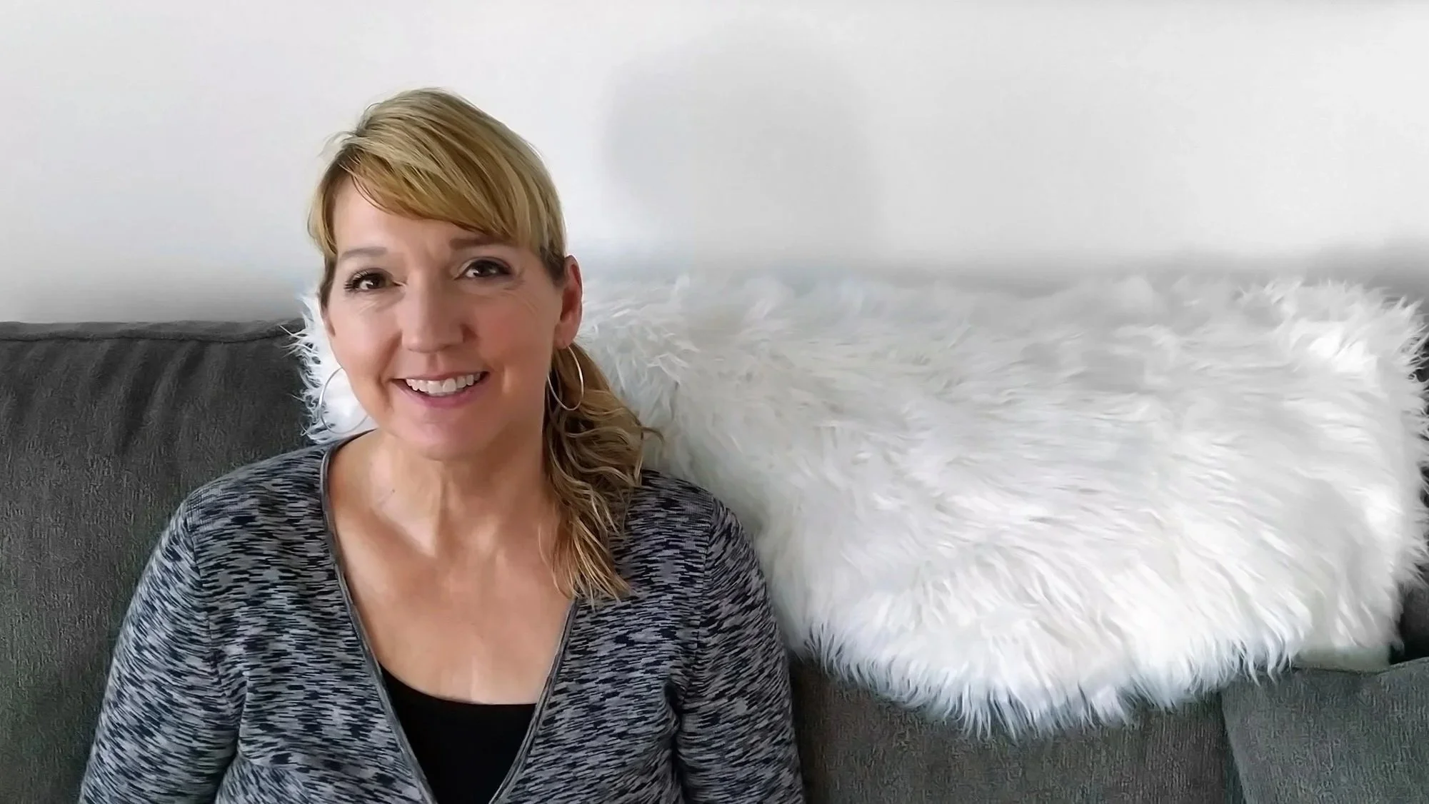 A smiling woman with blonde hair sitting on a gray couch with a white fluffy pillow in the background.