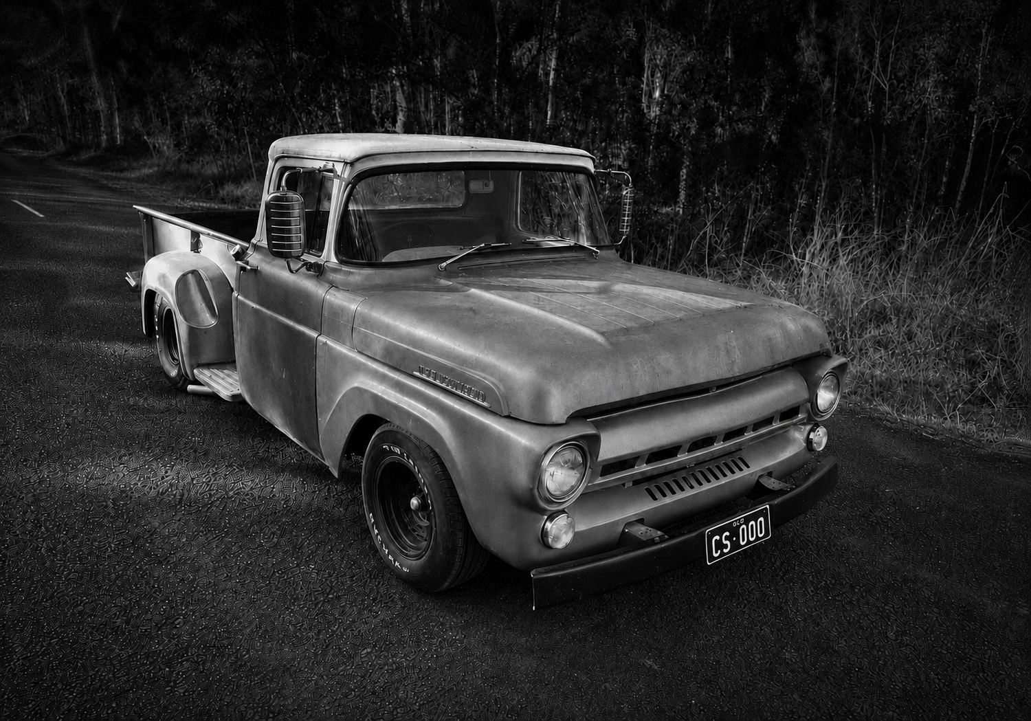A vintage pickup truck parked on a textured road with a forest background, shown in black and white.