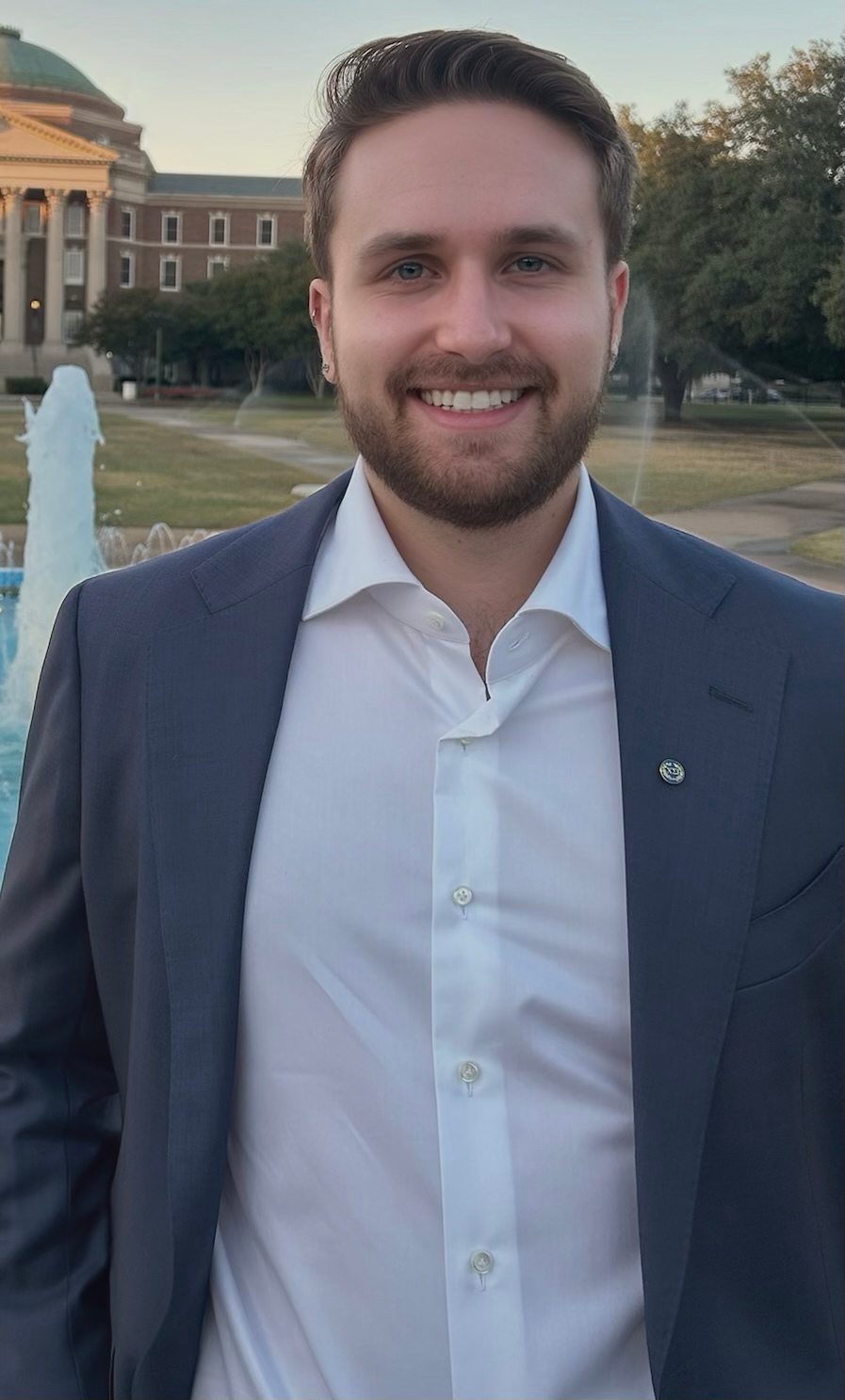 A smiling man with a beard and blue eyes, wearing a white shirt and a dark blue suit jacket, standing outdoors near a fountain with a historic government building in the background. It is likely late afternoon or early evening.