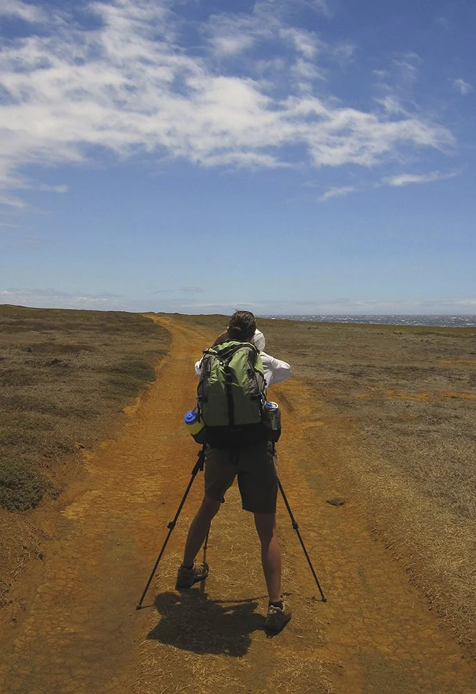 Person with a backpack hiking on a dirt trail in a flat, open landscape with blue sky and some clouds.