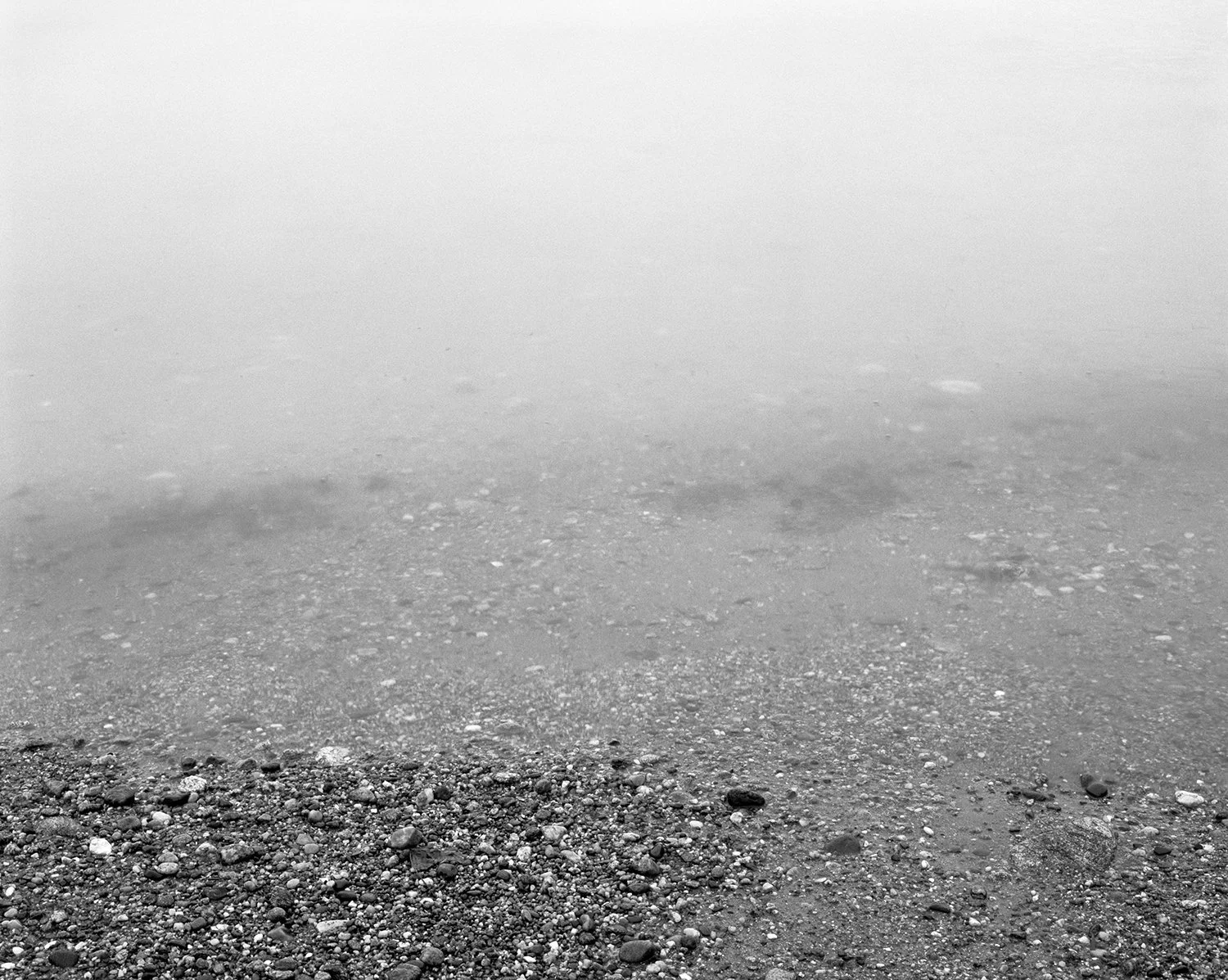 Black and white photo of a rocky shoreline with small pebbles and stones, foggy background obscuring the horizon.