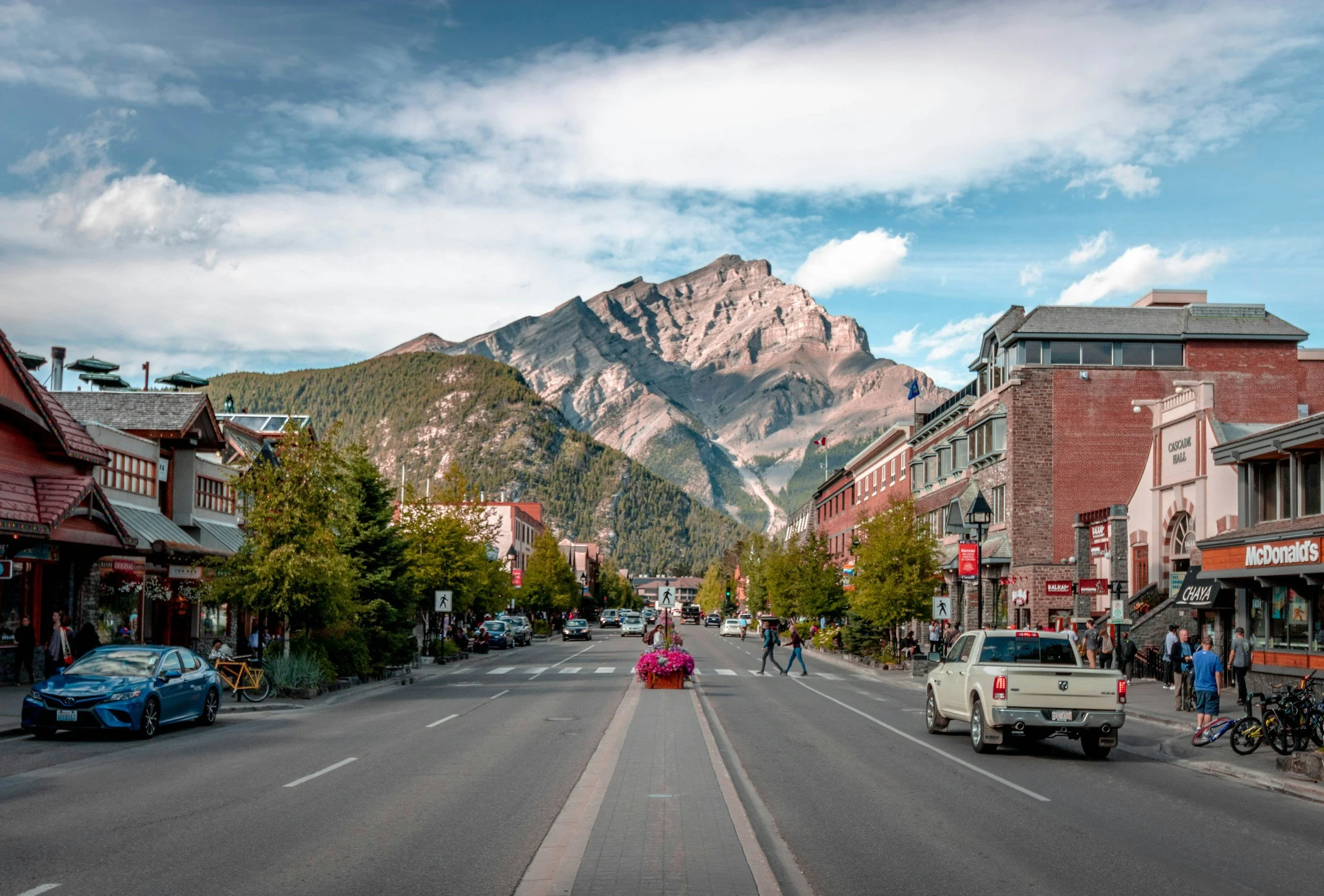 A bustling mountain town street with shops, pedestrians, cars, and trees, with a backdrop of rugged mountains and partly cloudy sky.