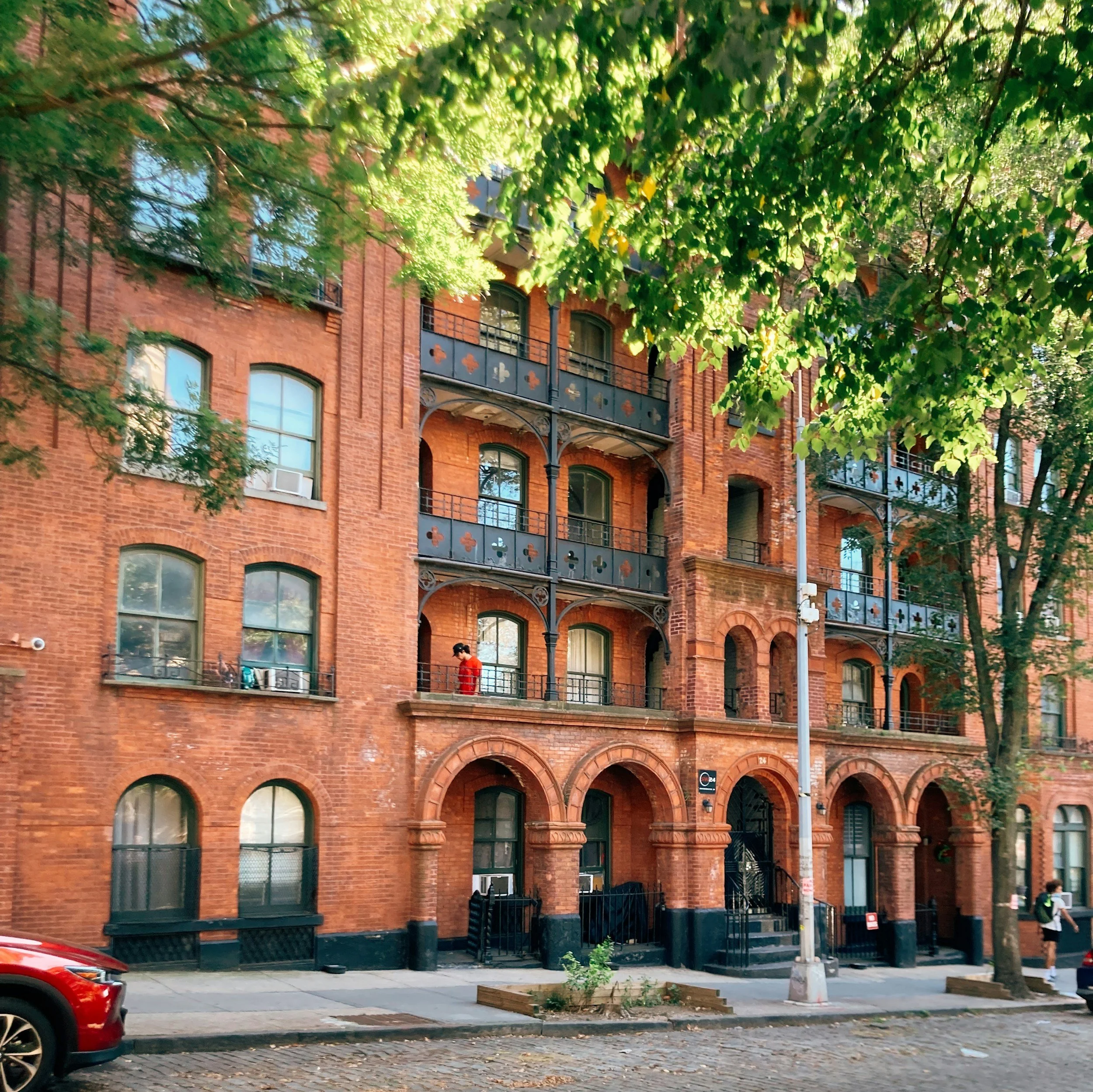 View of a red brick residential building with balconies, large windows, and decorative details, partially obscured by leafy green trees in an urban setting.