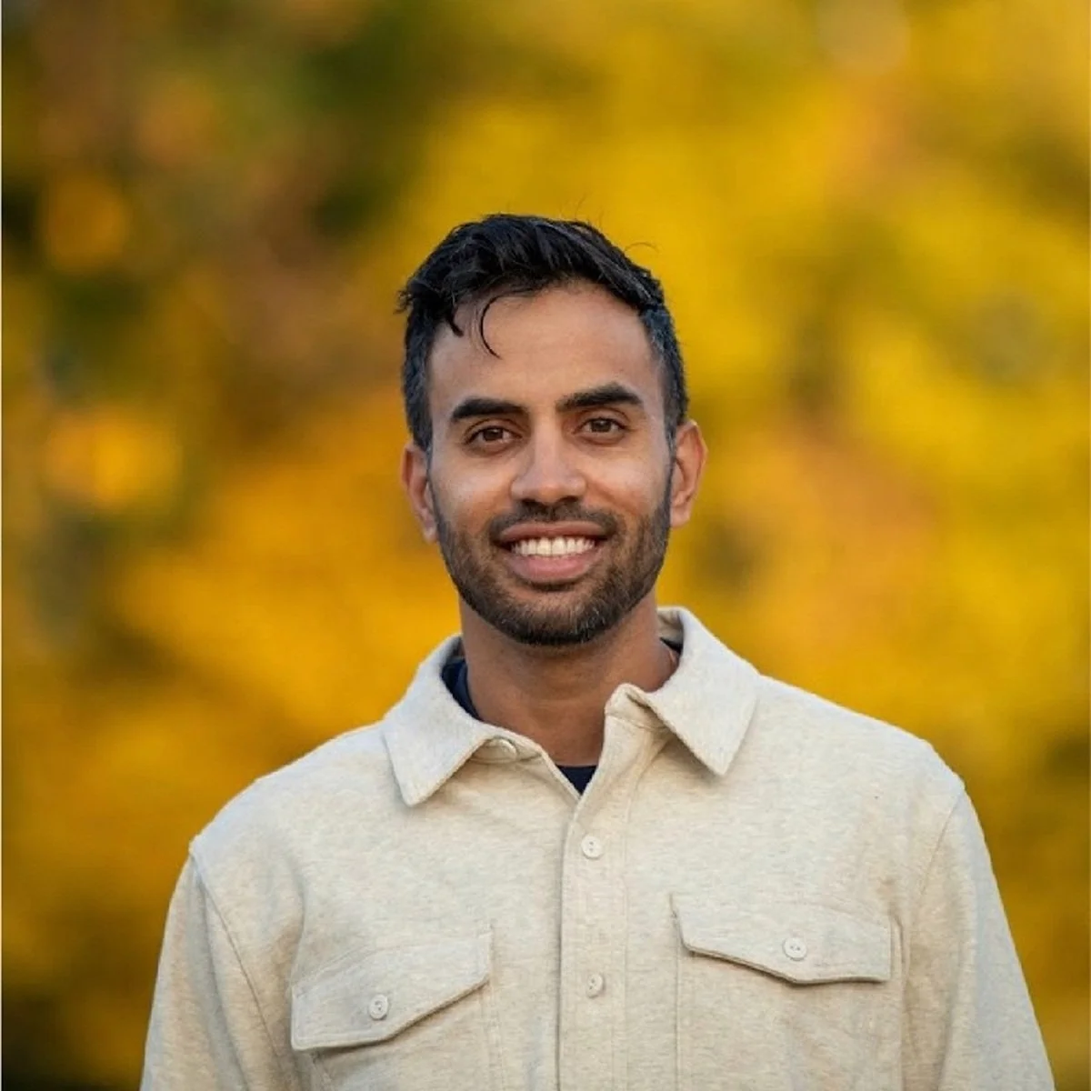A man smiling outdoors with a background of yellow autumn leaves.