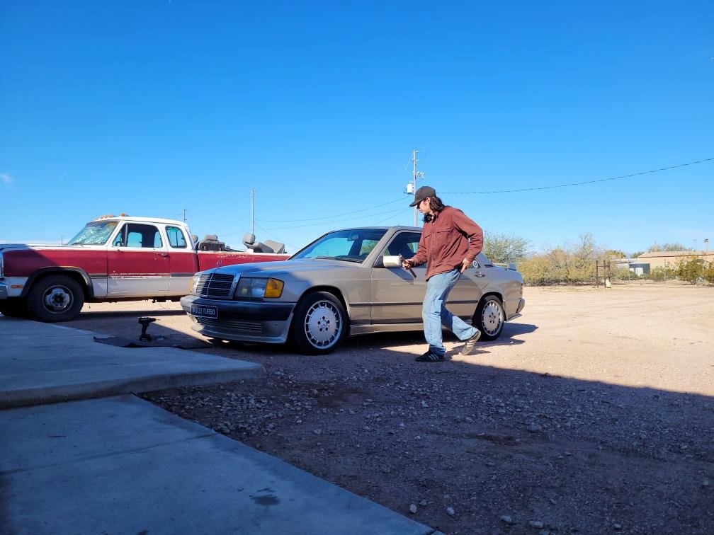 A man with long hair and a hat, wearing a brown jacket and jeans, is standing next to a silver car, looking at his phone, in a dirt lot with a blue sky overhead. An old two-tone pickup truck is parked nearby.