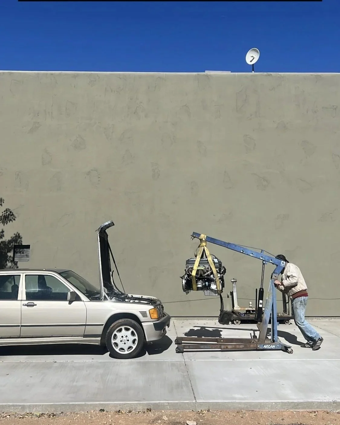 A man operating a crane to lift a removed engine from a silver car parked on the street, with a beige wall and satellite dish in the background.