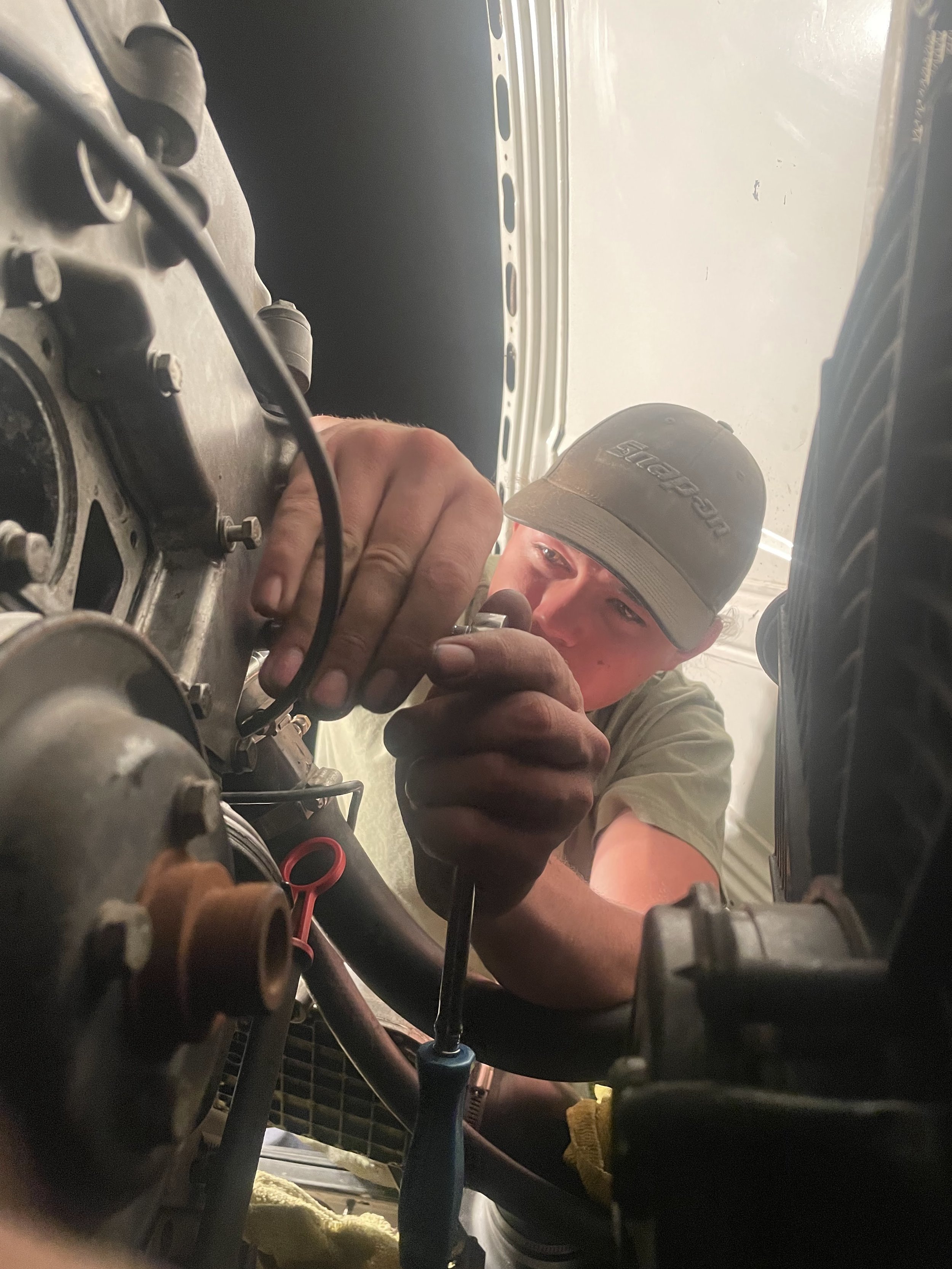 Man working on a mechanical component inside a vehicle engine compartment, wearing a cap, with tools nearby.