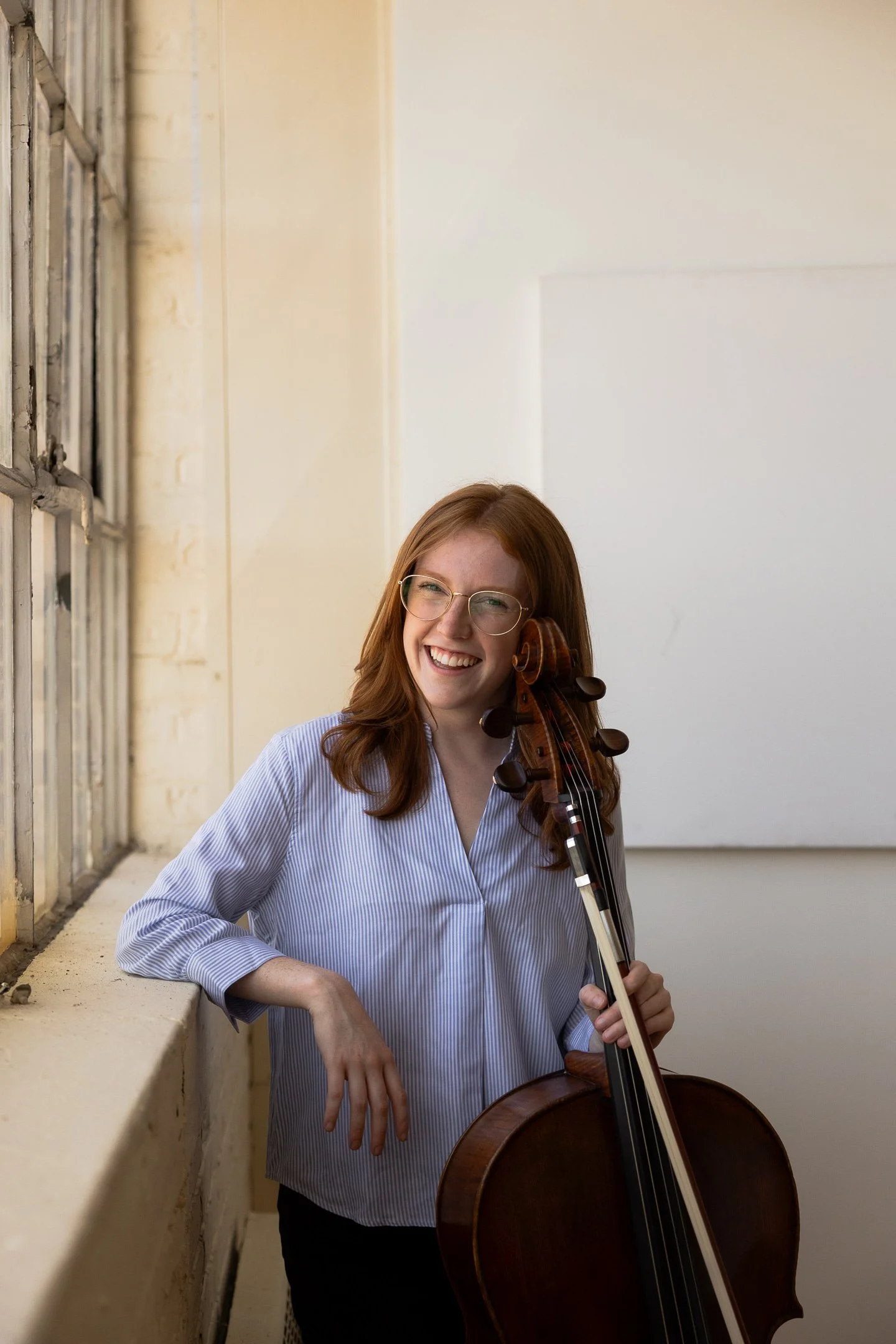 A young woman with red hair, glasses, and a striped blue shirt smiling while holding a cello and a bow, standing against a white wall and a windowed wall with an outdoor light.