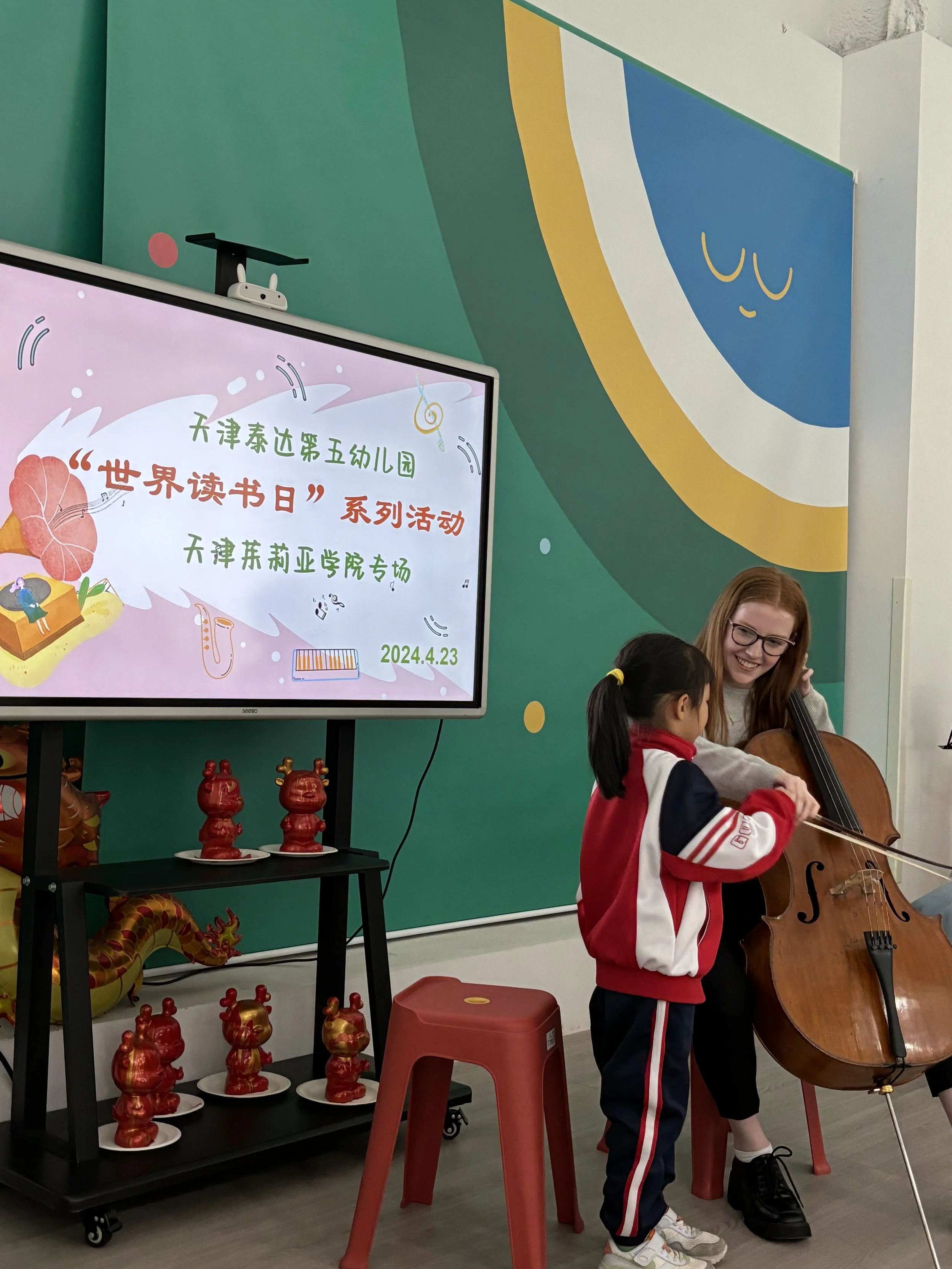 A young girl and a woman playing a cello together at a music event. The girl is wearing a red and white jacket and black pants, while the woman is smiling and has long red hair and glasses. Behind them, there is a colorful wall with a blue and yellow design, and a large screen displaying Chinese characters and illustrations for a reading event.