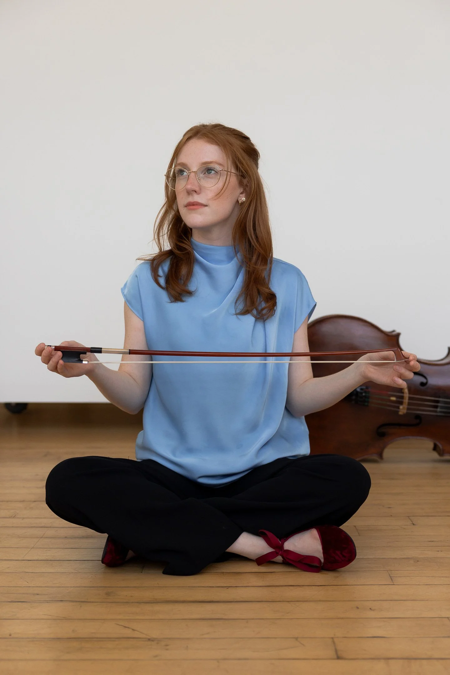 A young woman with red hair and glasses sitting cross-legged on a wooden floor, playing a violin.
