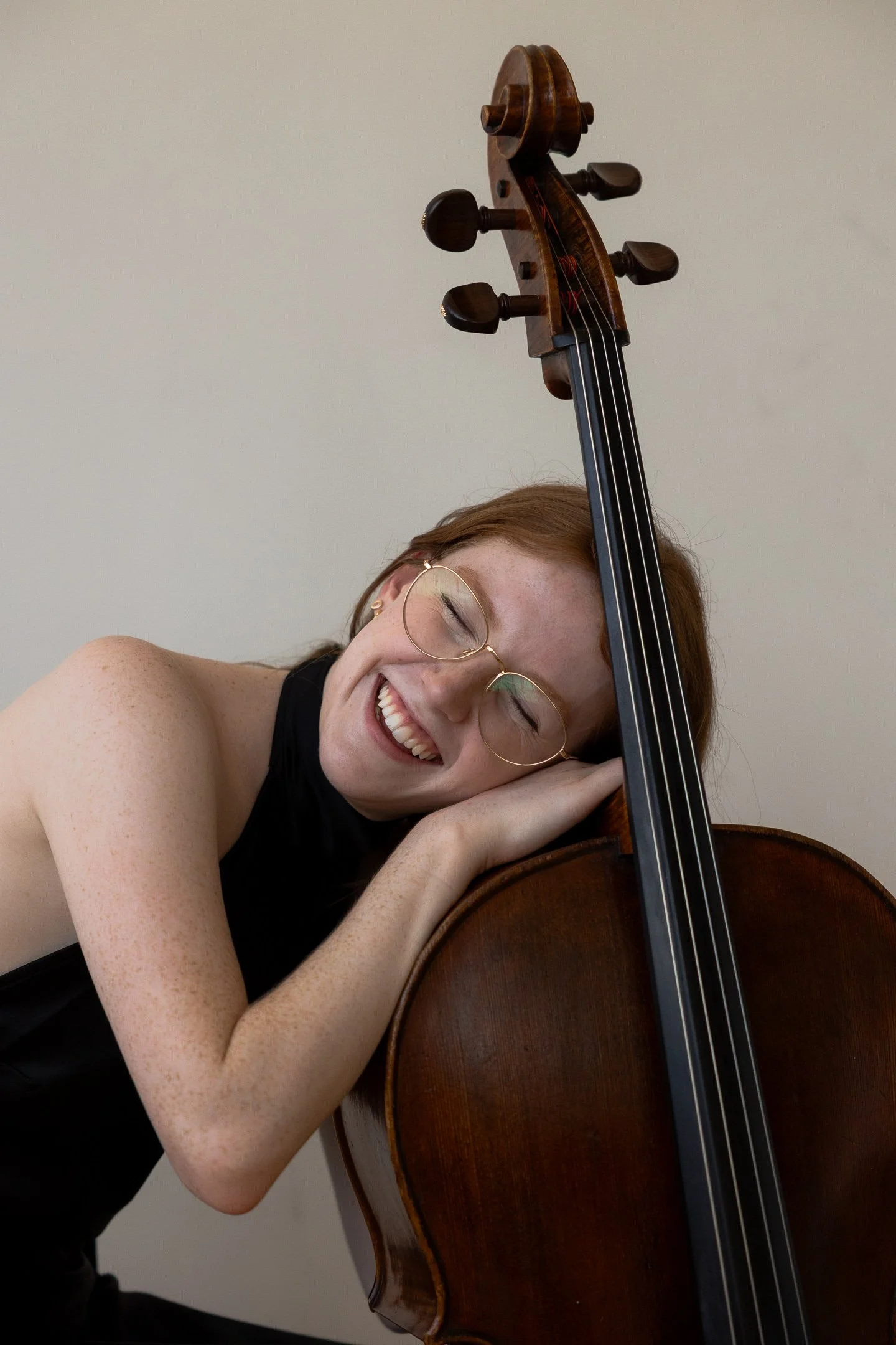 A woman with glasses and freckles smiling with her eyes closed, resting her head on a cello's body and hugging the instrument.