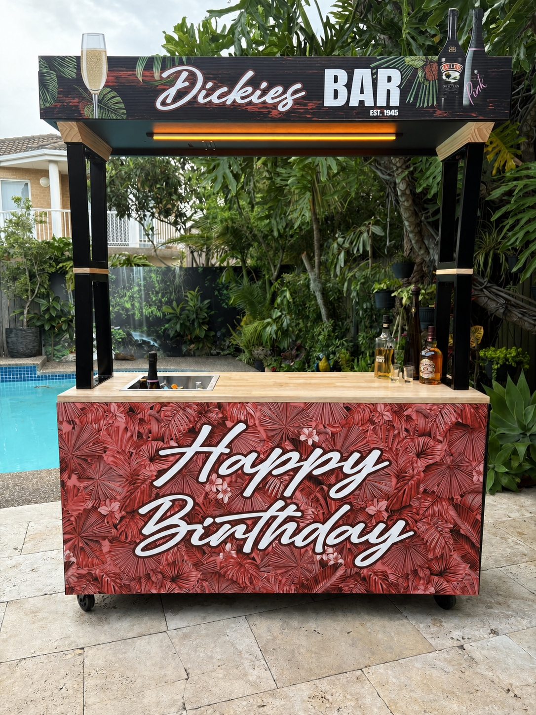 Outdoor portable bar with custom graphics 'Happy Birthday' & 'Dickies BAR' with illustrations of a champagne glass and bottles. The bar has bottles of alcohol on the counter.