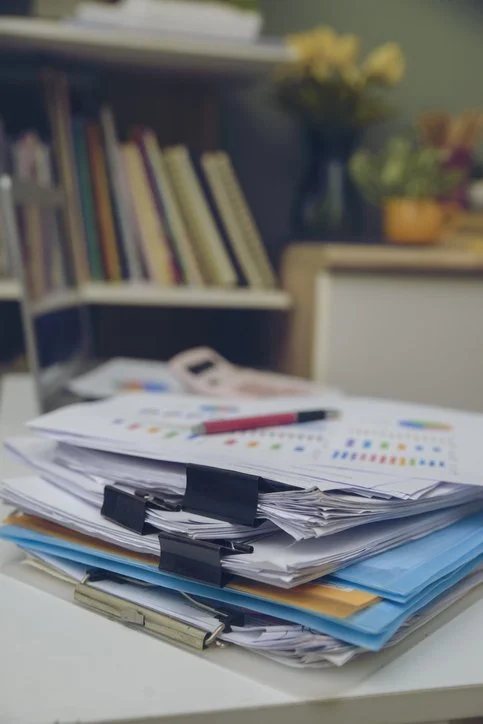 Piled papers with colored graphs and a pen on top of a desk, with a bookshelf and flowers in the background.