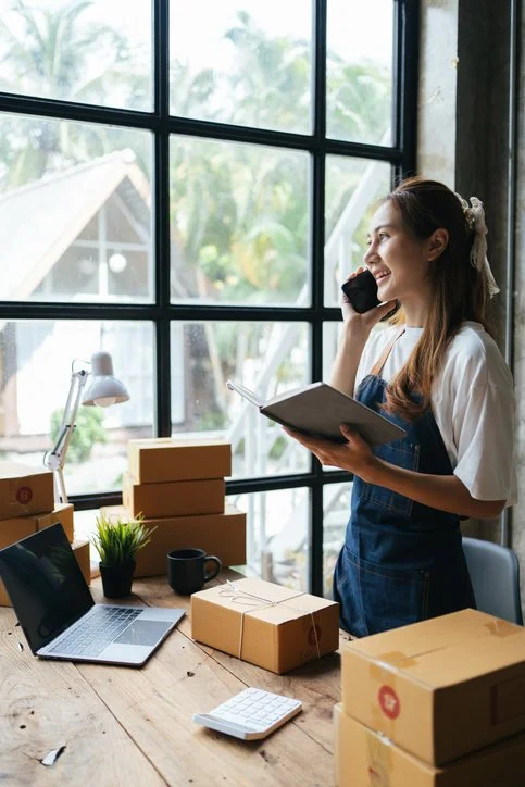 A woman stands at a wooden desk with a laptop, boxes, a plant, mug, and papers, talking on her phone inside a bright room with large windows showing an outdoor view.